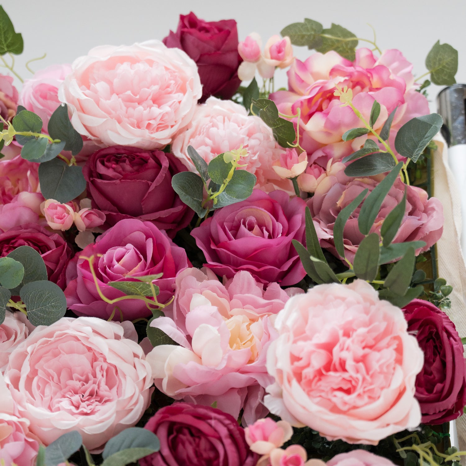 A close - up of artificial flowers: soft pink peonies, deep - pink roses, and tiny buds, all surrounded by green eucalyptus leaves. The lush, layered petals and varied hues create a romantic, realistic arrangement—perfect for decorative displays, adding a touch of elegance and charm.
