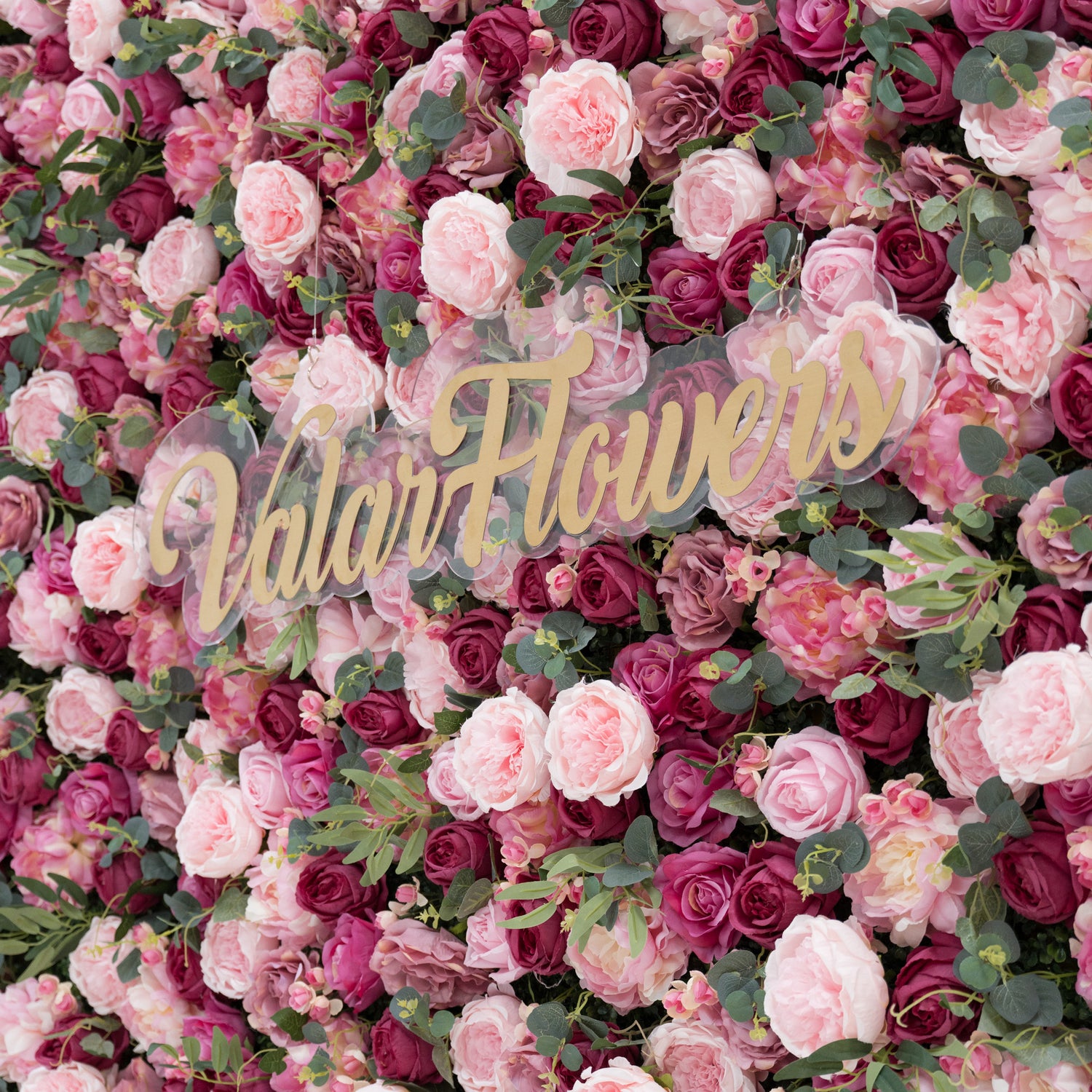 A lush artificial flower wall filled with pink and deep - pink roses, plus green foliage. A clear acrylic sign with gold “ValarFlowers” lettering stands out, creating an elegant, branded backdrop—ideal for events, blending natural charm and sophisticated branding.