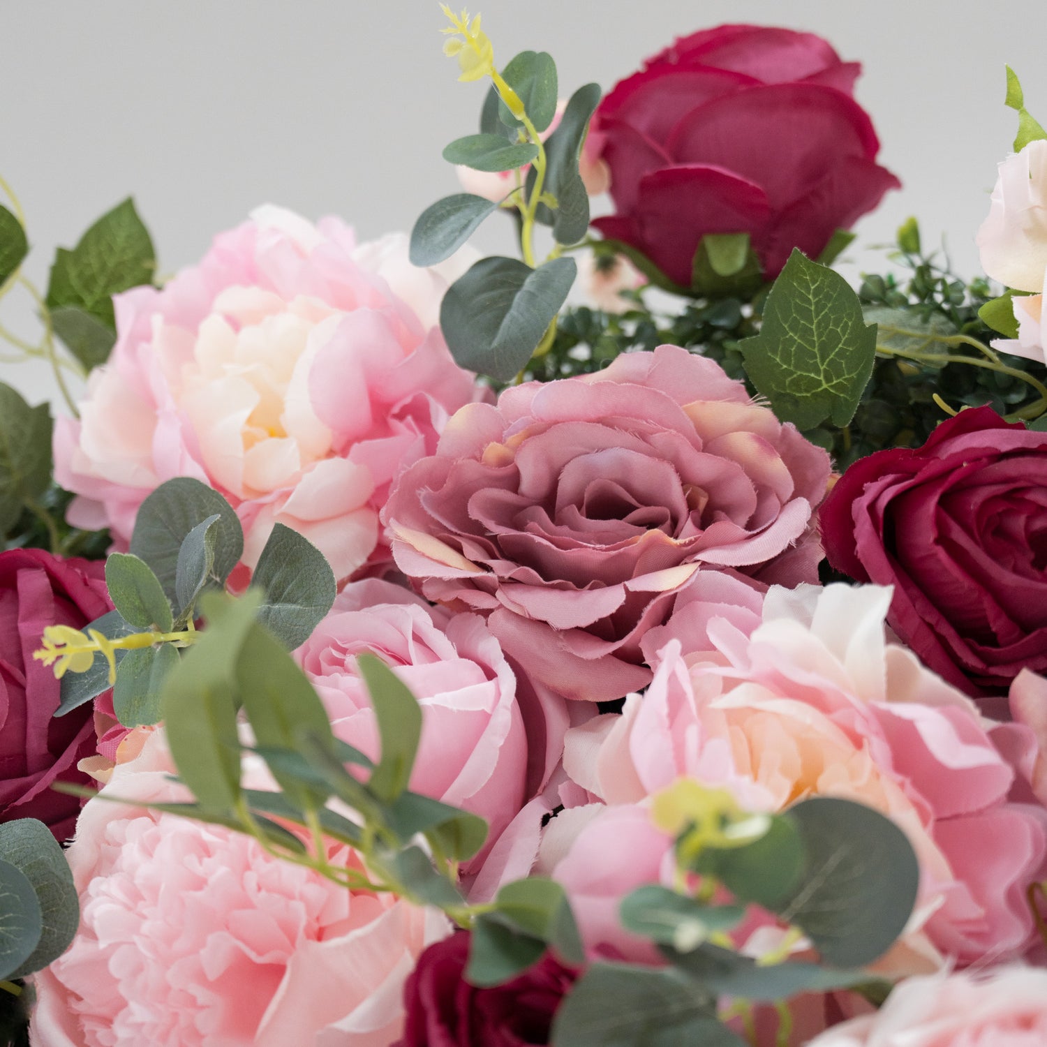 A close - up of artificial flowers: soft pink peonies, delicate light - pink roses, and deep - red blooms, all interspersed with green foliage. The lush, layered petals and varied hues create a romantic, realistic arrangement—perfect for decorative displays, adding charm and elegance.