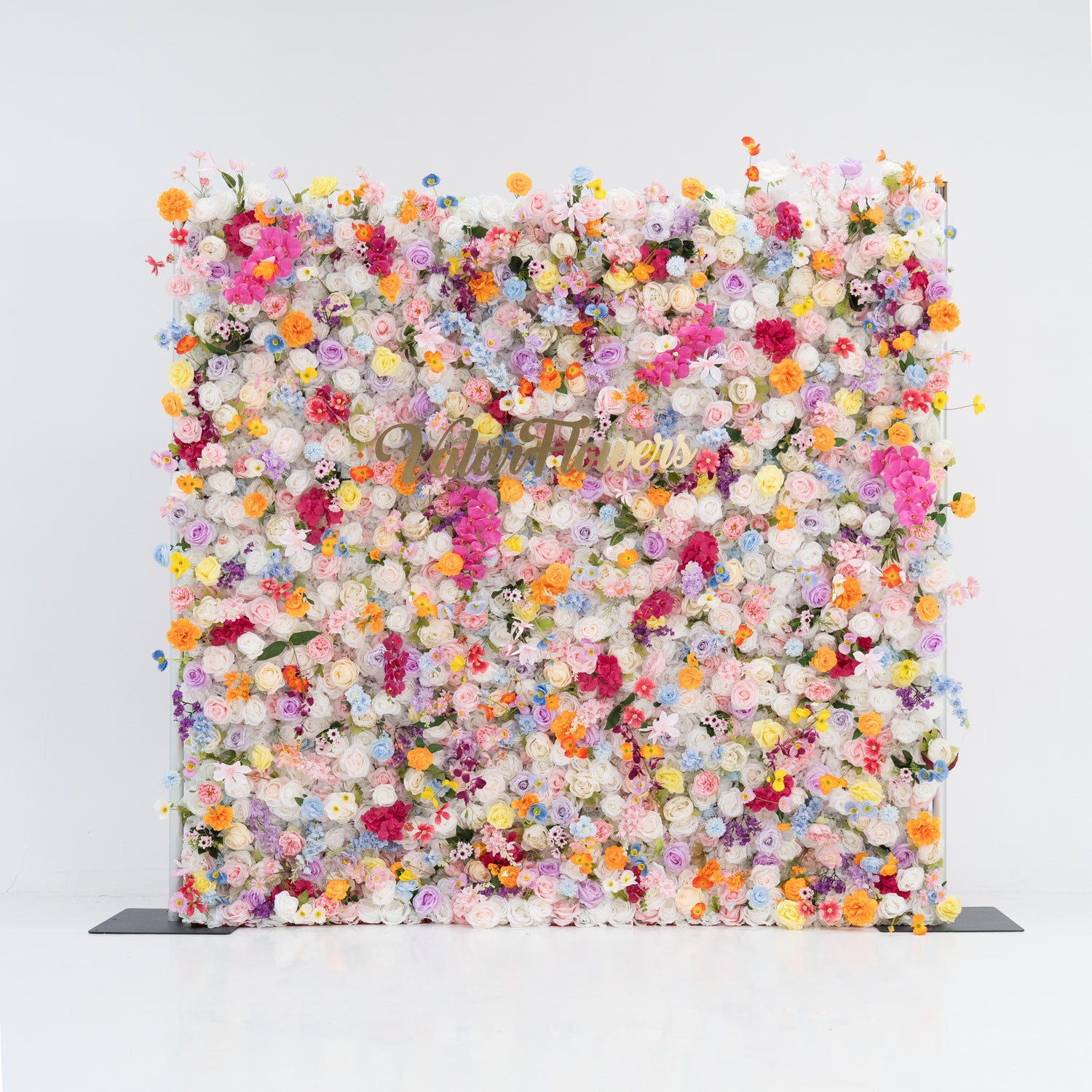 A large, square floral wall bursts with a vibrant mix of colorful artificial flowers—pink, yellow, orange, purple, and more. Dense clusters create a lush, lively display. With the “Valor Flowers” sign in the center, set against a plain white background and floor, it exudes a cheerful, festive vibe, perfect for events like weddings, parties, or as a decorative photo backdrop.