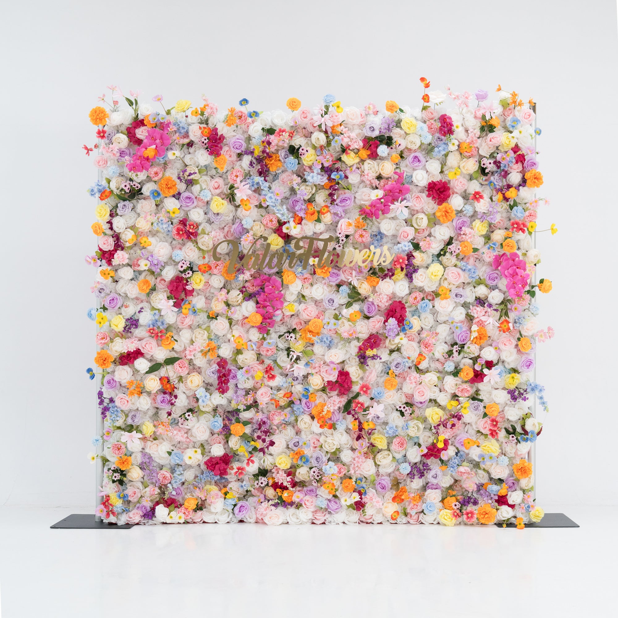 A large, square floral wall bursts with a vibrant mix of colorful artificial flowers—pink, yellow, orange, purple, and more. Dense clusters create a lush, lively display. With the “Valor Flowers” sign in the center, set against a plain white background and floor, it exudes a cheerful, festive vibe, perfect for events like weddings, parties, or as a decorative photo backdrop.