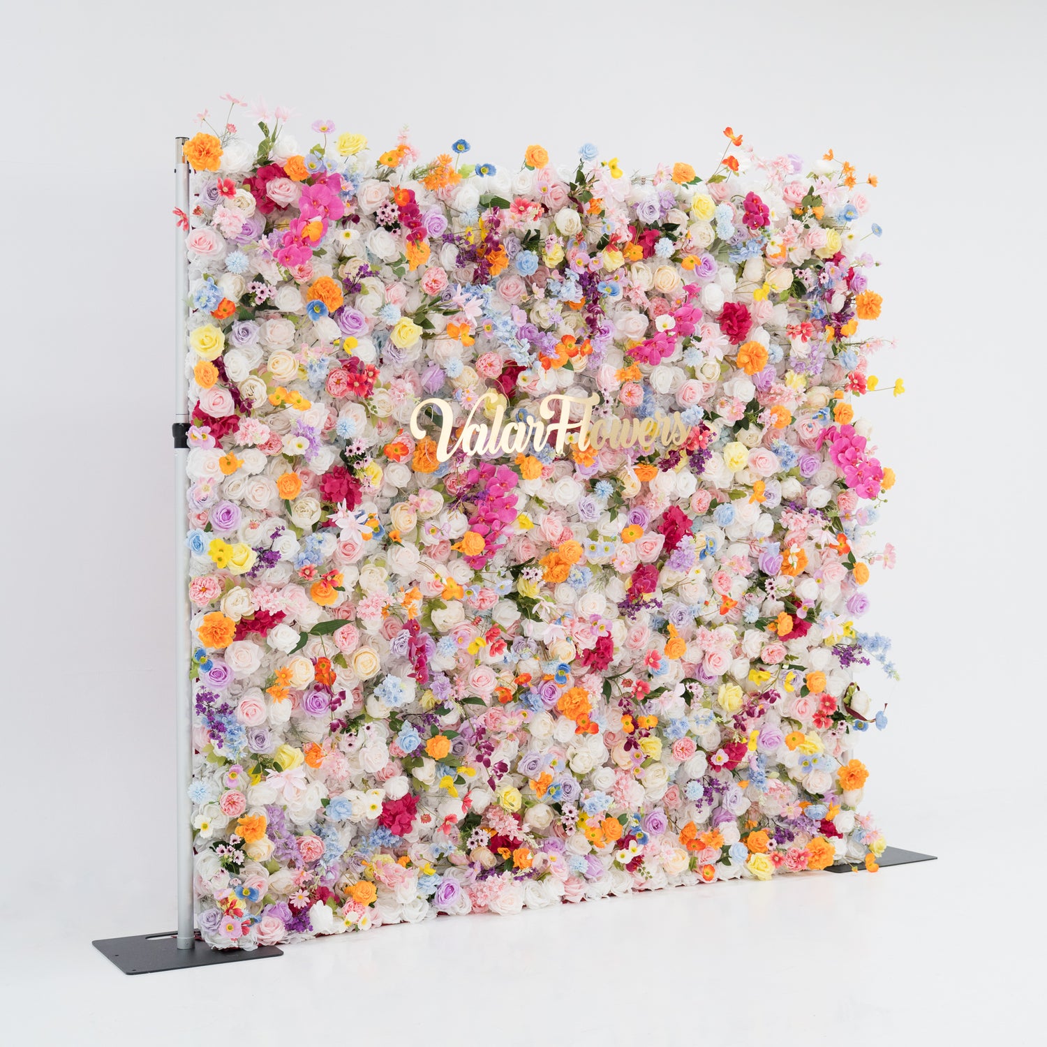 A large floral wall brims with a vibrant mix of colorful artificial flowers—pink, yellow, orange, purple, and more. Dense clusters create a lush, lively display. With the “Valar Flowers” sign in the center, set against a plain white background and floor, it exudes a cheerful, festive vibe, perfect for events like weddings, parties, or as a decorative photo backdrop.