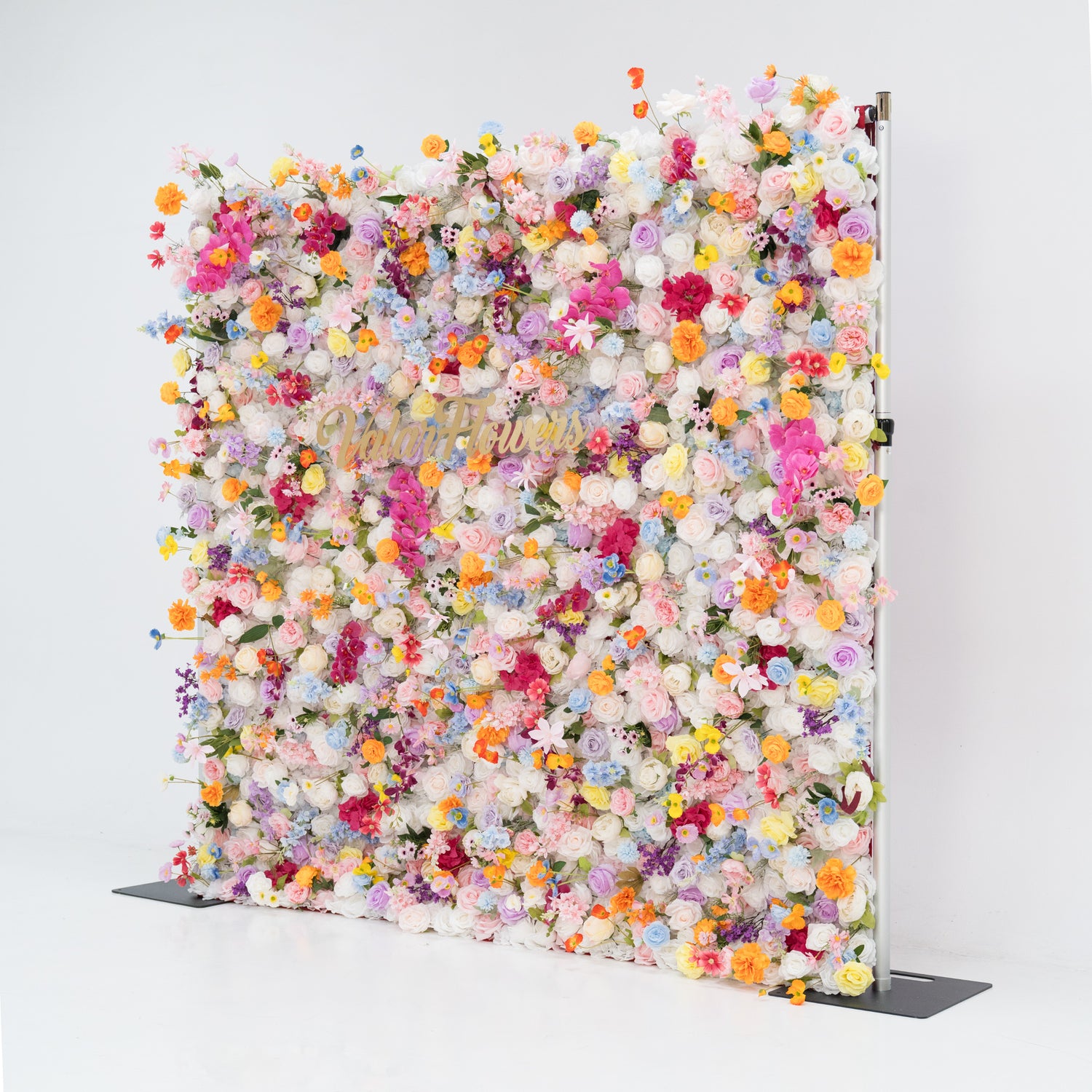 A large floral wall overflows with a vibrant mix of colorful artificial flowers—pink, yellow, orange, purple, and more. Dense clusters create a lush, lively display. Featuring the “Valar Flowers” sign, set against a plain white background and floor, it exudes a cheerful, festive vibe, perfect for events like weddings, parties, or as a decorative photo backdrop.