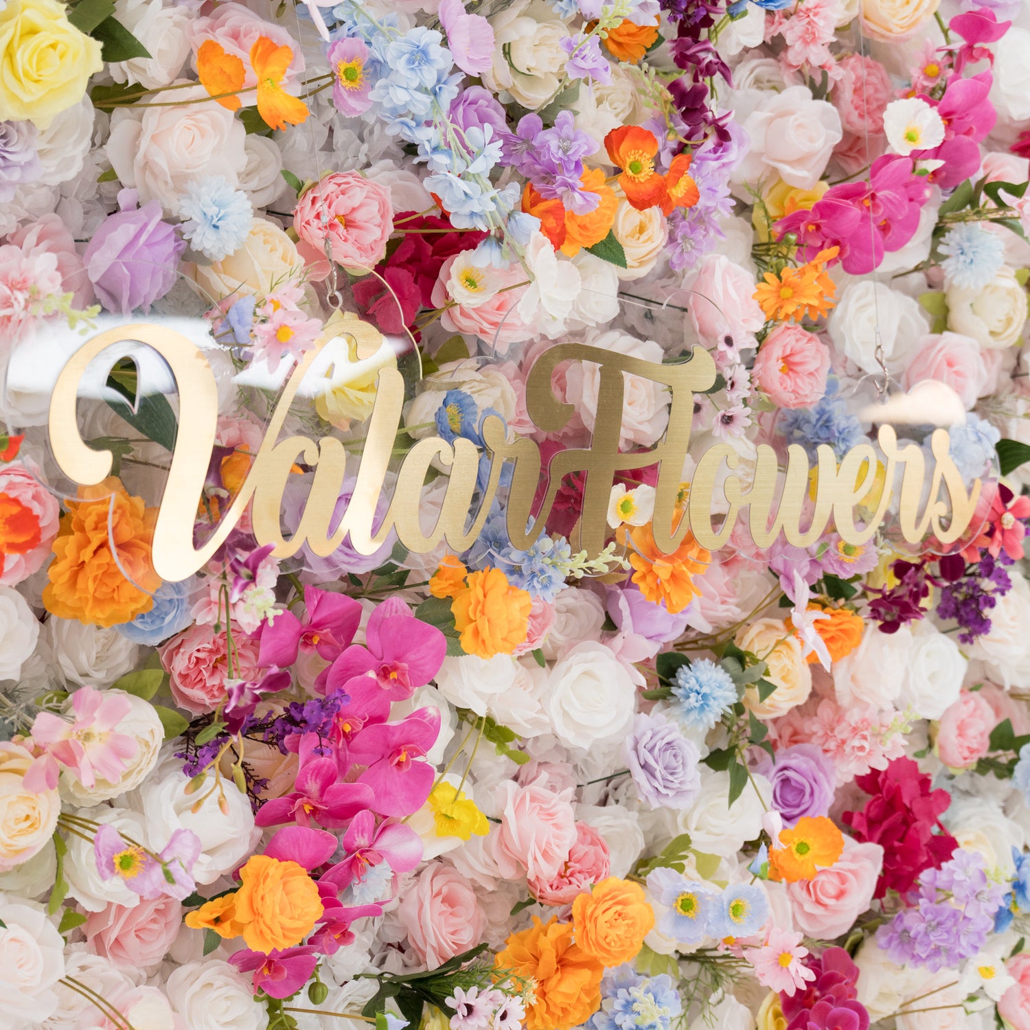 A vibrant floral wall is filled with a rich mix of artificial flowers in colors like pink, orange, purple, and white. At the center, a golden “Valar Flowers” sign stands out. The dense, diverse blooms create a lively, cheerful scene, perfect for decorative backdrops at events such as weddings or parties, exuding a festive and elegant vibe.