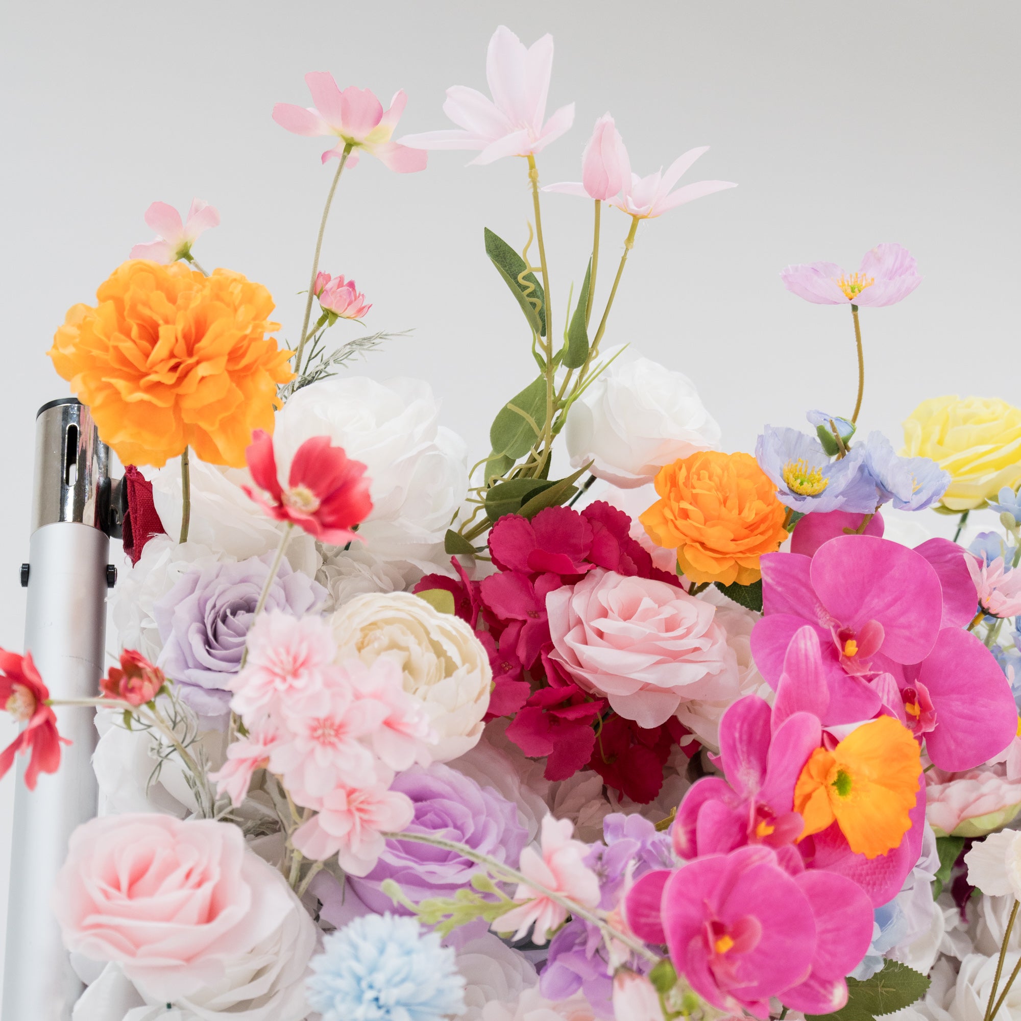 A vibrant arrangement of artificial flowers in a rich array of colors—orange, pink, purple, red, and more—creates a lively display. Various floral types, with layered petals and delicate details, contribute to a lush, cheerful scene. Set against a plain light - colored background, it exudes a festive and elegant vibe, perfect for decorative purposes at events like weddings or parties.