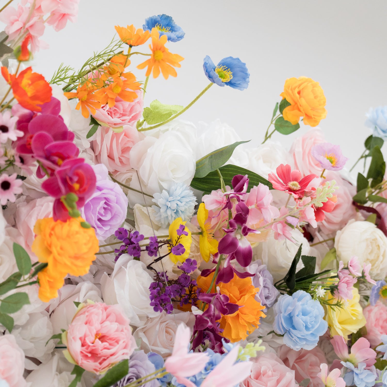 A vibrant arrangement of artificial flowers bursts with colors—orange, pink, purple, blue, and more. Various floral types, with layered petals and delicate details, create a lush, cheerful display. Set against a plain light - colored background, the dense, diverse blooms exude a festive and elegant vibe, perfect for decorative backdrops at events like weddings or parties.