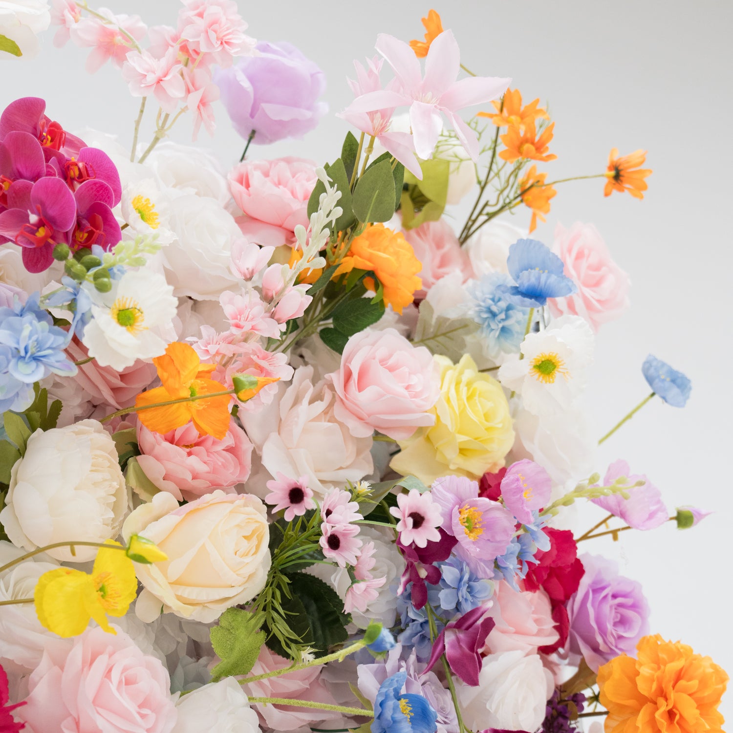 A vibrant arrangement of artificial flowers showcases a rich palette of colors—pink, orange, yellow, blue, purple, and more. Various floral types, with layered petals and delicate details, create a lush, cheerful display. Set against a plain light - colored background, the dense, diverse blooms exude a festive and elegant vibe, perfect for decorative backdrops at events like weddings or parties.