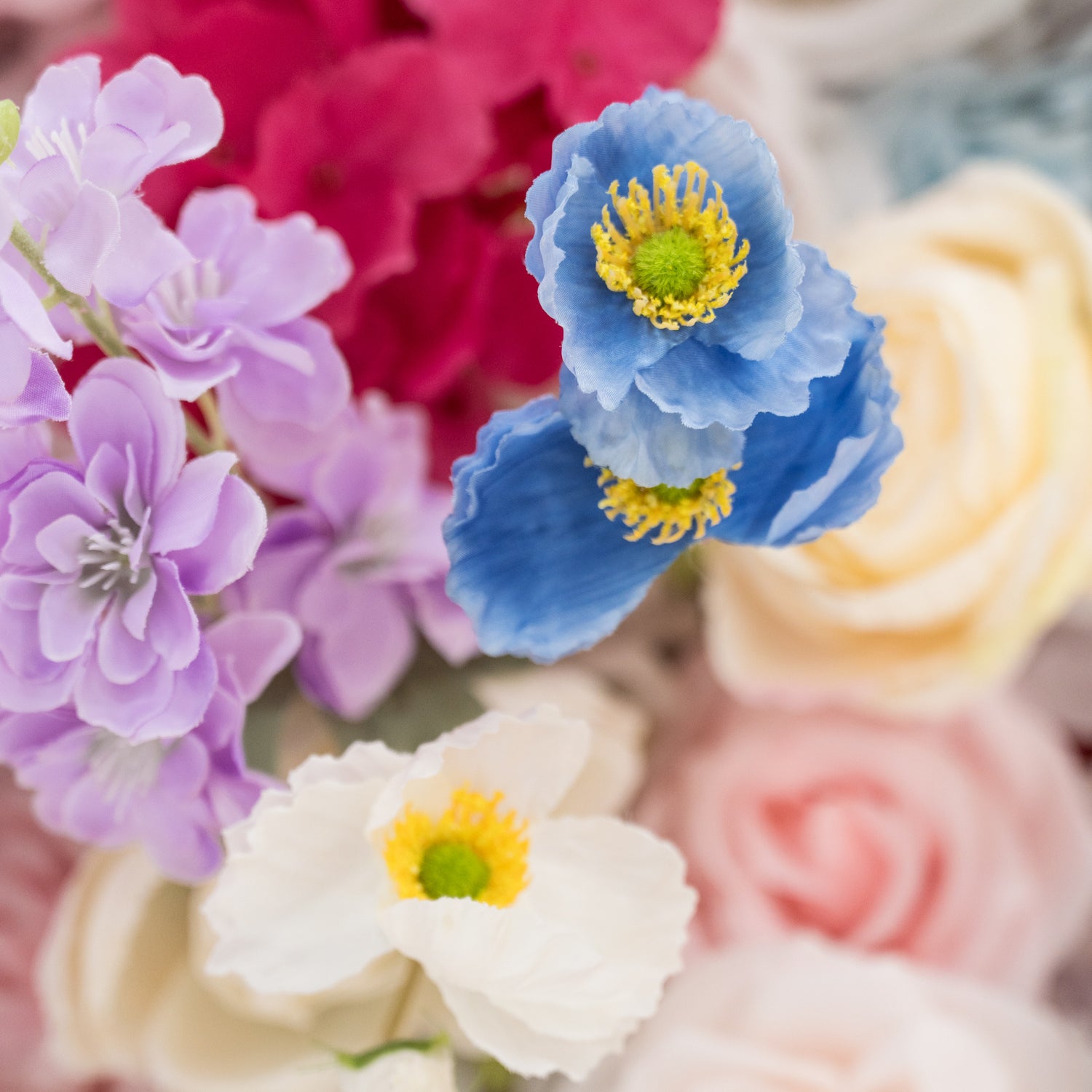 A close - up of artificial flowers features a lovely mix of colors. There are blue flowers with yellow centers, purple blooms, and a white flower with a yellow and green center. In the background, hints of pink and red flowers add depth. The petals have a soft, realistic texture, creating a delicate and charming floral arrangement, perfect for decorative purposes.