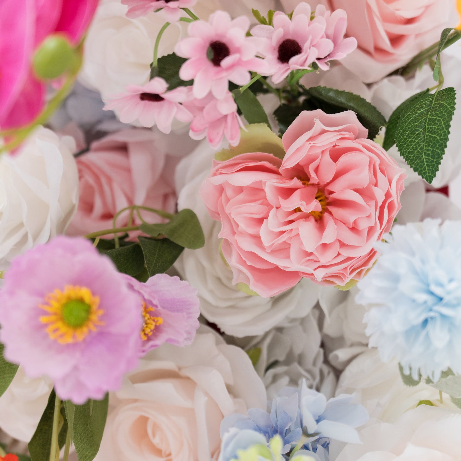 A vibrant arrangement of artificial flowers features a lovely mix of colors. There are soft pink roses, delicate pink daisies with dark centers, a prominent pink bloom with layered petals, and a purple flower with a yellow center. Alongside, there are white roses and light blue blooms, all complemented by green leaves. The petals have a smooth, realistic texture, creating a charming and elegant floral display, perfect for decorative purposes.