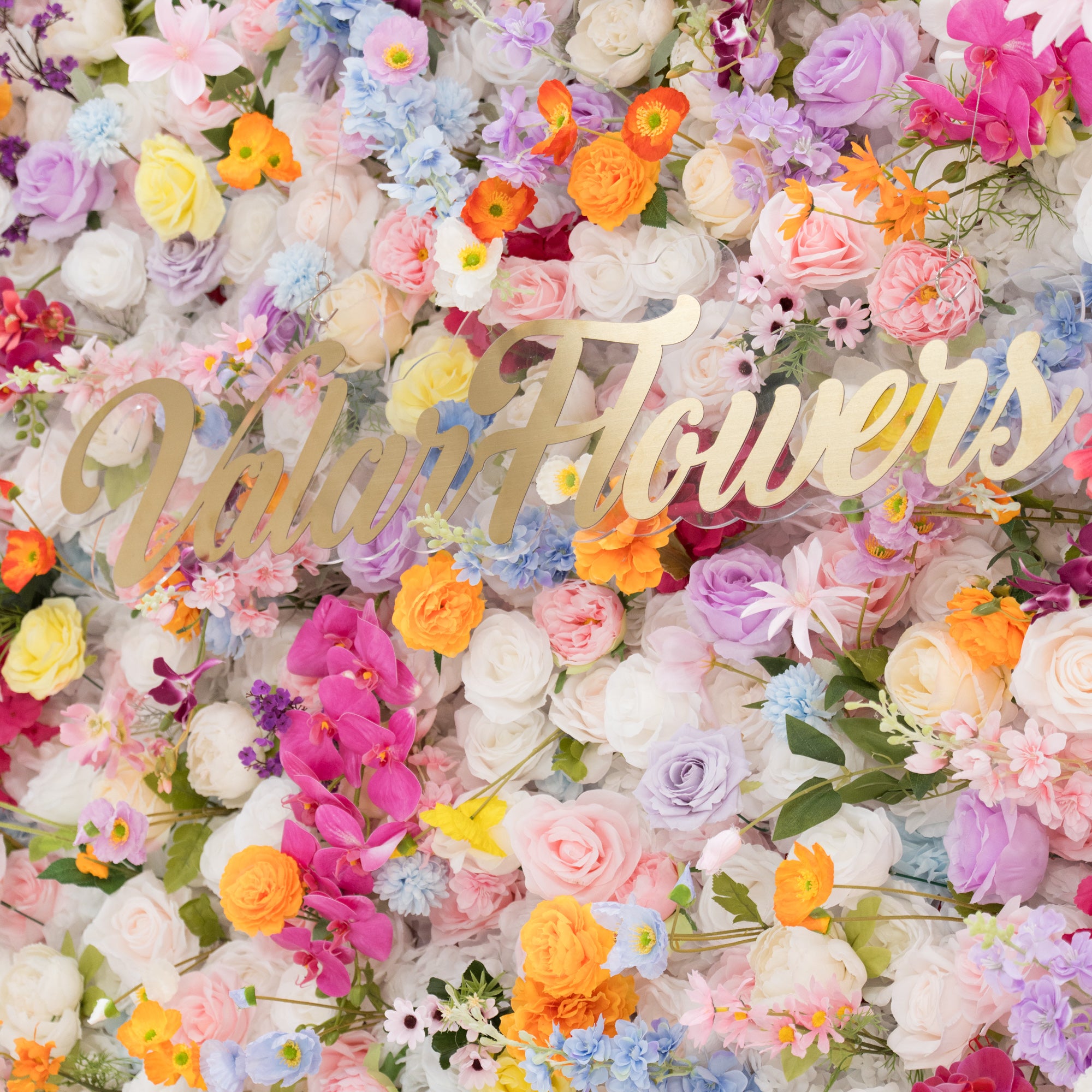 A vibrant floral wall is filled with a rich mix of artificial flowers in colors like pink, orange, purple, and white. At the center, a golden “Valar Flowers” sign stands out. The dense, diverse blooms create a lively, cheerful scene, perfect for decorative backdrops at events such as weddings or parties, exuding a festive and elegant vibe.