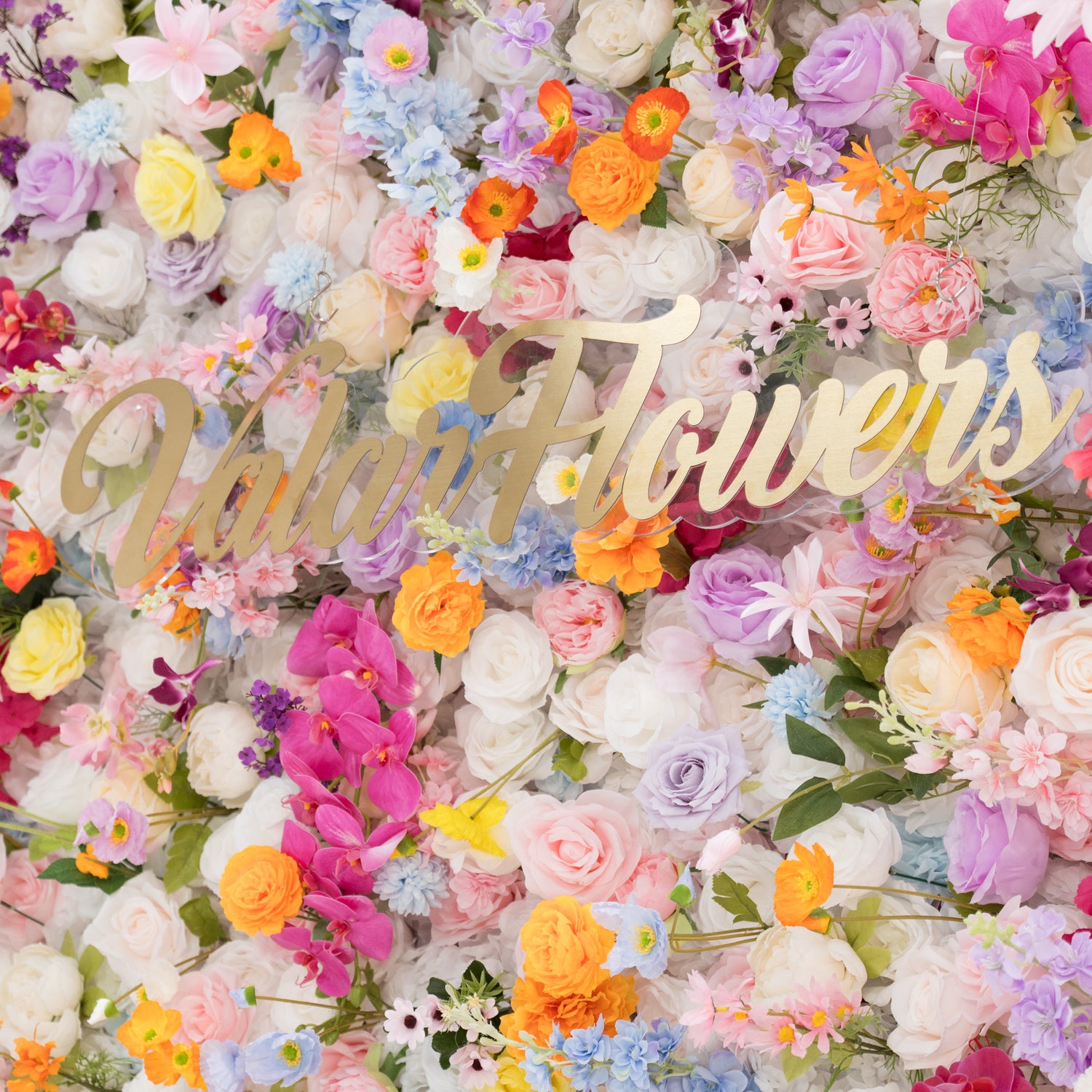 A vibrant floral wall is filled with a rich mix of artificial flowers in colors like pink, orange, purple, and white. At the center, a golden “Valar Flowers” sign stands out. The dense, diverse blooms create a lively, cheerful scene, perfect for decorative backdrops at events such as weddings or parties, exuding a festive and elegant vibe.