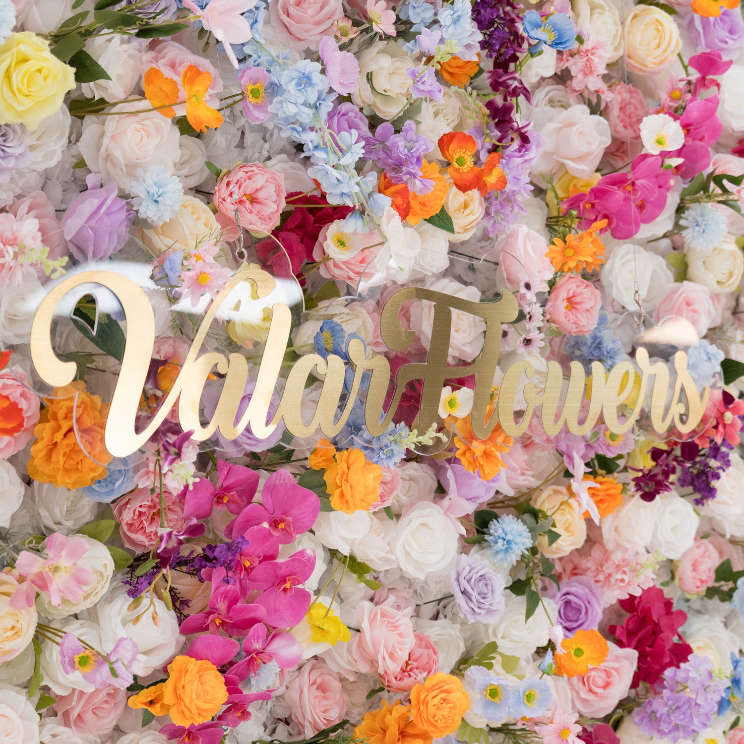 A vibrant floral wall is filled with a rich mix of artificial flowers in colors like pink, orange, purple, and white. At the center, a golden “Valar Flowers” sign stands out. The dense, diverse blooms create a lively, cheerful scene, perfect for decorative backdrops at events such as weddings or parties, exuding a festive and elegant vibe.