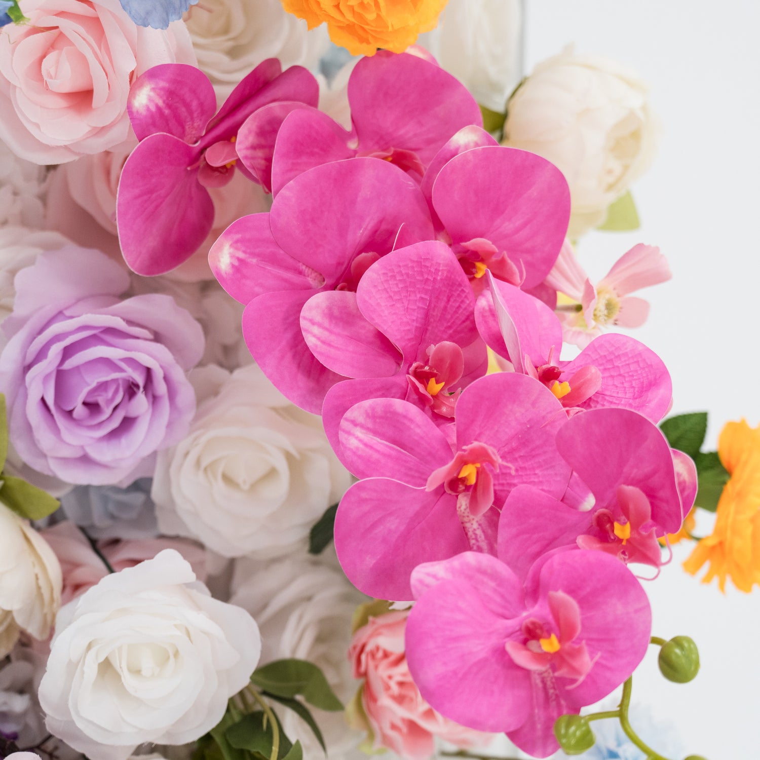 This image presents a lush arrangement of artificial flowers. Prominently, there are vibrant pink orchids with delicate petals and bright yellow centers. Surrounding them are roses in shades of white, light purple, and soft pink, along with hints of other blooms like orange flowers. The flowers, with their realistic textures, create a visually appealing and elegant display, ideal for decorative purposes in settings such as events or interiors.