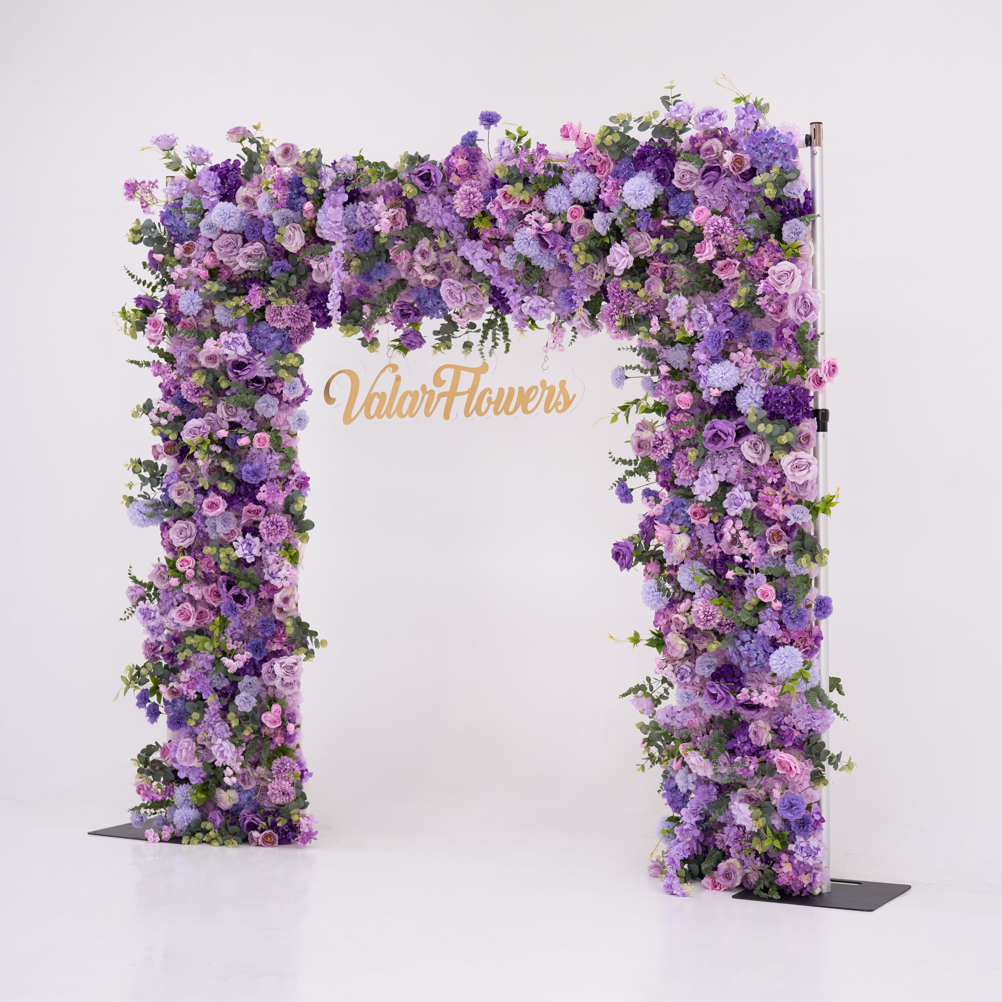This image showcases a stunning floral arch by Valar Flowers, set against a plain white indoor background. It features a dense arrangement of artificial flowers in various shades of purple, lavender, and pink, creating an elegant, nature-inspired display. The brand’s name is elegantly integrated in gold, making it perfect for adding a luxurious, sophisticated element to events or interiors, highlighting Valar Flowers’ craftsmanship in eye-catching decor.