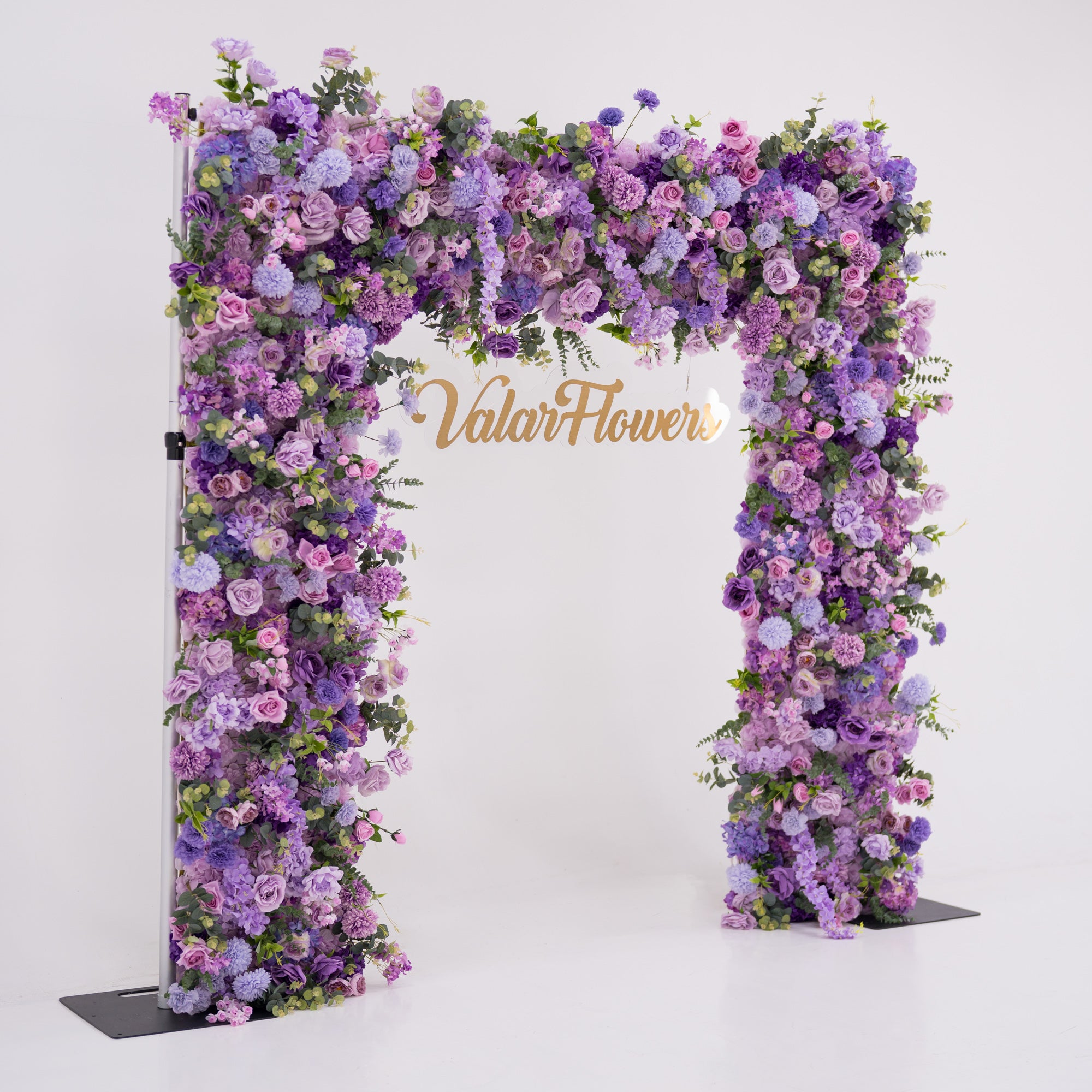 This image showcases a stunning floral arch by Valar Flowers, set against a plain white indoor background. It features a dense arrangement of artificial flowers in various shades of purple, lavender, and pink, creating an elegant, nature-inspired display. The brand’s name is elegantly integrated in gold, making it perfect for adding a luxurious, sophisticated element to events or interiors, highlighting Valar Flowers’ craftsmanship in eye-catching decor.