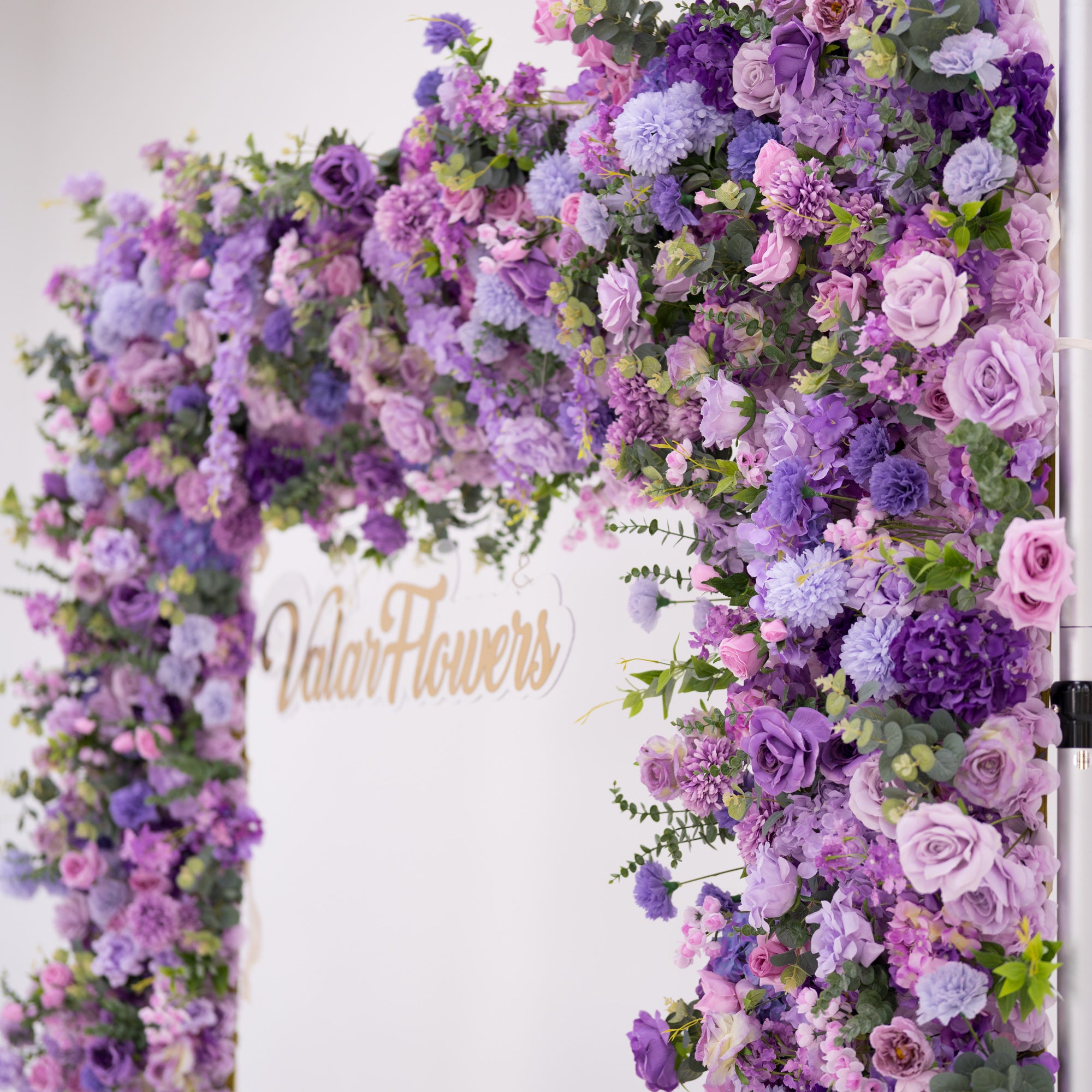 This image showcases a vibrant section of a floral arch by Valar Flowers, featuring a dense arrangement of artificial flowers in various shades of purple, lavender, and pink. The textured display includes roses, hydrangeas, and other blooms, complemented by green foliage. The brand’s name is elegantly integrated in gold, exuding elegance and perfect for adding a luxurious, nature-inspired element to events or interiors.