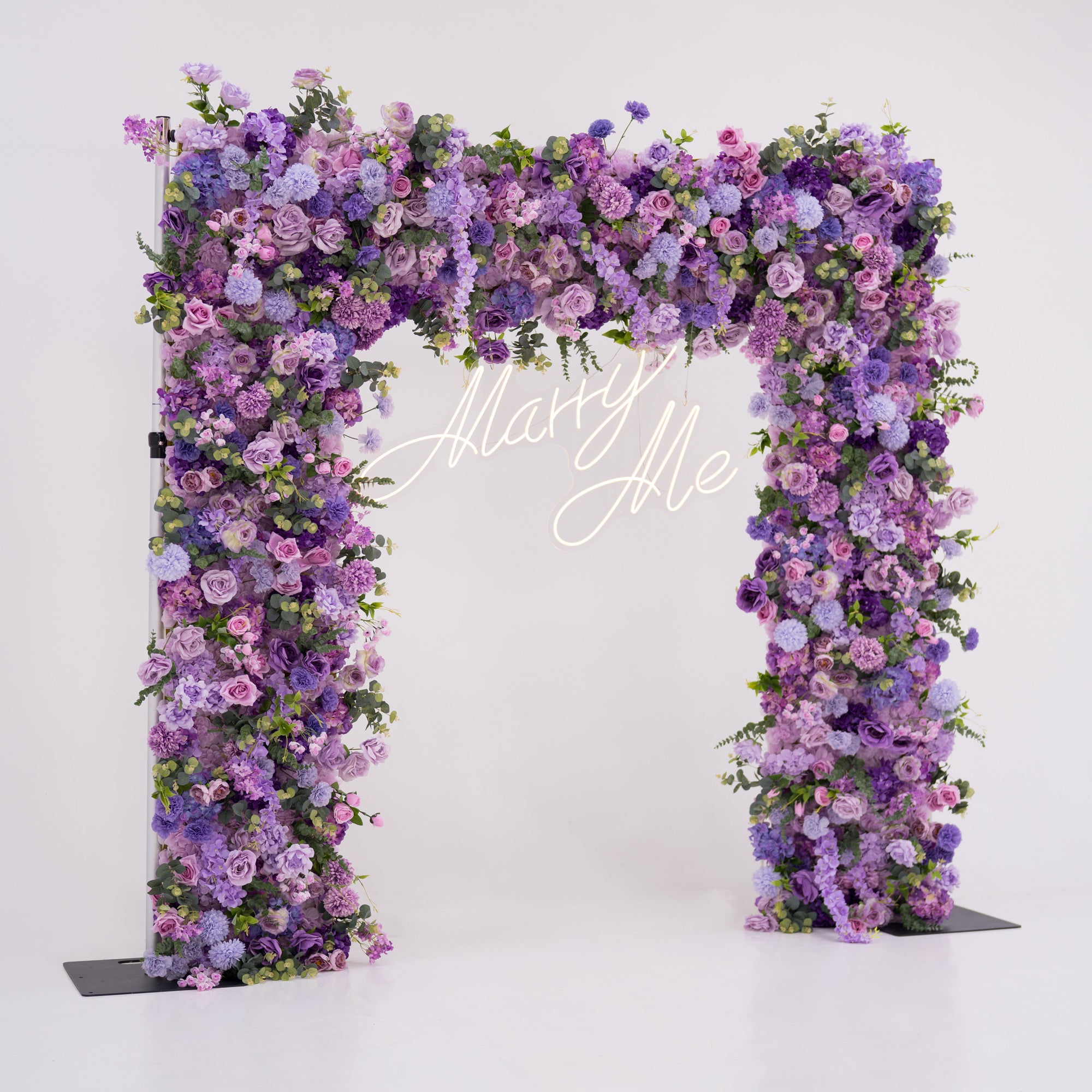 This image showcases a stunning floral arch by Valar Flowers, set against a plain white indoor background. It features a dense arrangement of artificial flowers in various shades of purple, lavender, and pink, with a “Marry Me” neon sign at the center. Exuding romance and elegance, it’s perfect for proposals or wedding events, highlighting Valar Flowers’ craftsmanship in creating luxurious, eye-catching decor.