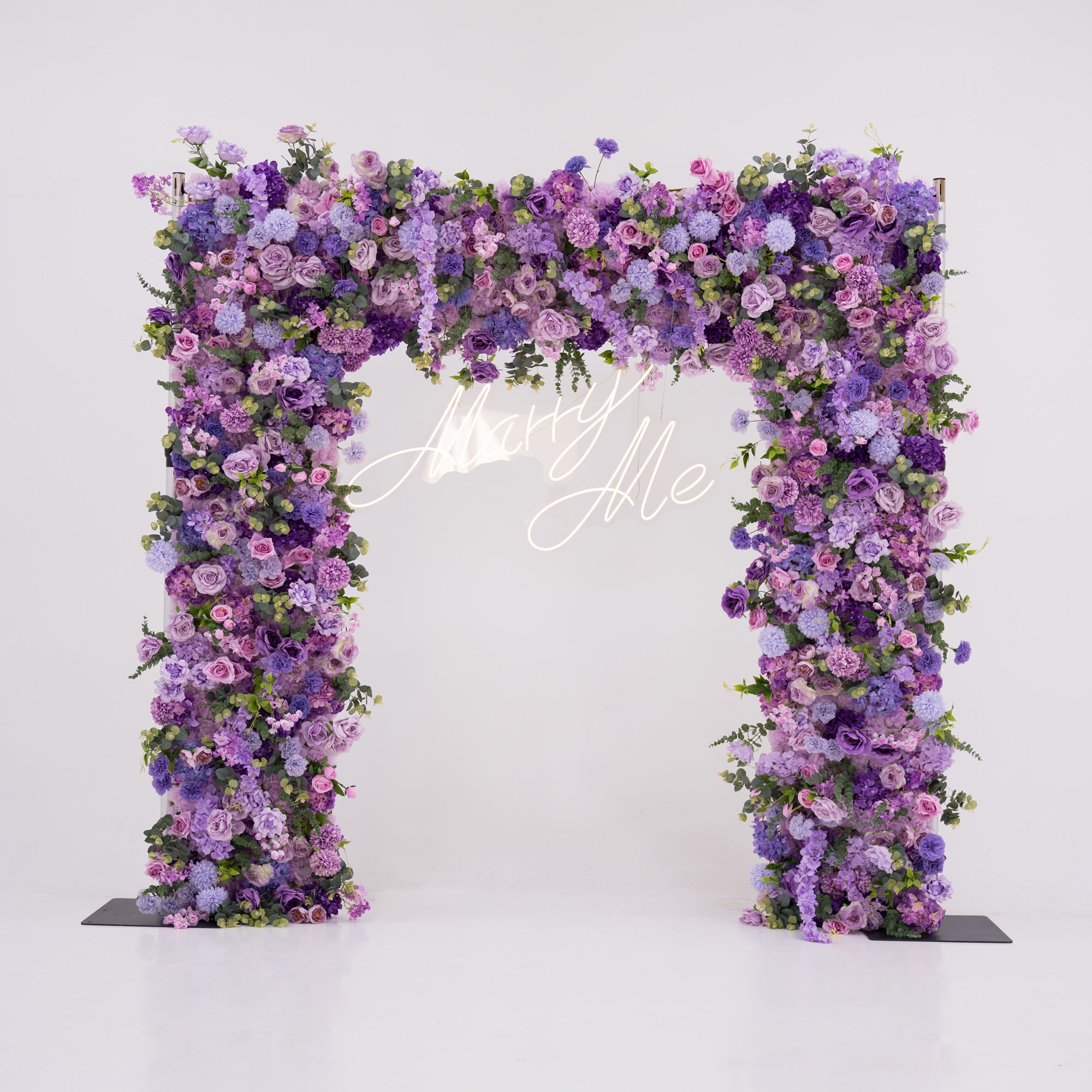 This image showcases a stunning floral arch by Valar Flowers, set against a plain white indoor background. It features a dense arrangement of artificial flowers in various shades of purple, lavender, and pink, with a “Marry Me” neon sign at the center. Exuding romance and elegance, it’s perfect for proposals or wedding events, highlighting Valar Flowers’ craftsmanship in creating luxurious, eye-catching decor.