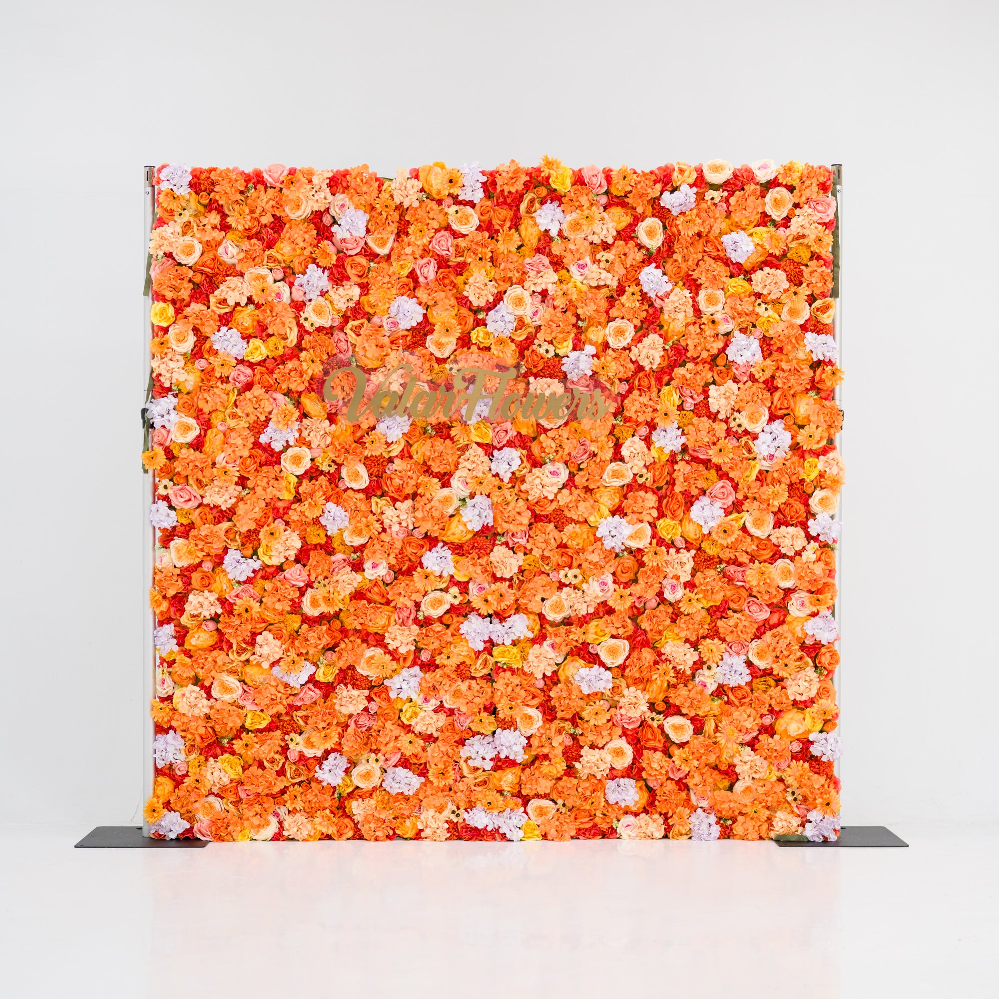 This image showcases a vibrant floral backdrop by Valar Flowers, featuring a dense arrangement of artificial flowers in warm shades of orange, peach, and hints of pink. The textured display exudes energy and warmth, with the brand’s name elegantly integrated. Set against a plain white indoor background, it’s perfect for adding a bold, nature-inspired element to events or interiors.