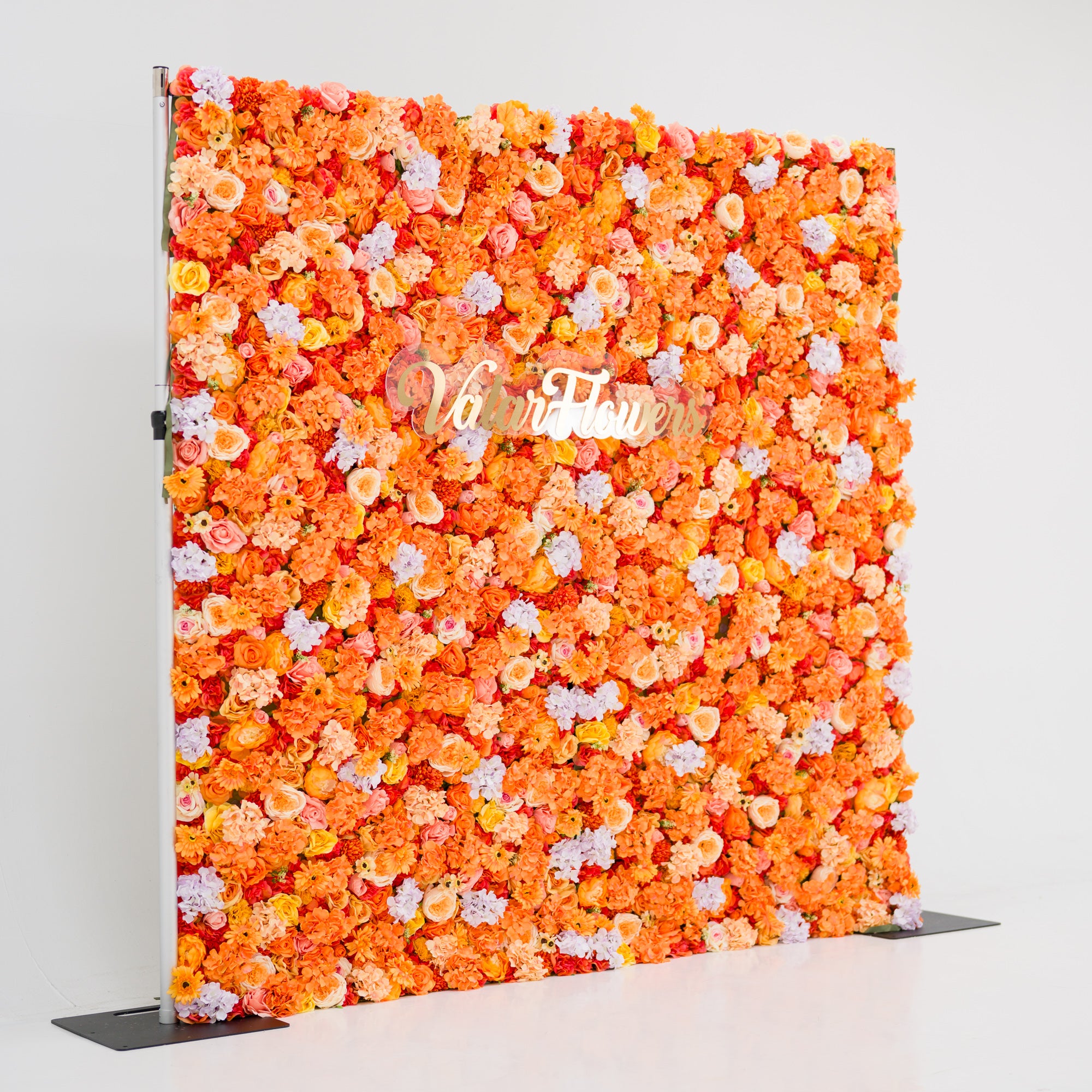 This image showcases a vibrant floral backdrop by Valar Flowers, featuring a dense arrangement of artificial flowers in warm shades of orange, peach, and hints of pink and purple. The textured display exudes energy and warmth, with the brand’s name elegantly integrated in gold. Set against a plain white indoor background, it’s perfect for adding a bold, nature-inspired element to events or interiors, highlighting Valar Flowers’ craftsmanship in creating eye-catching, sophisticated decor.