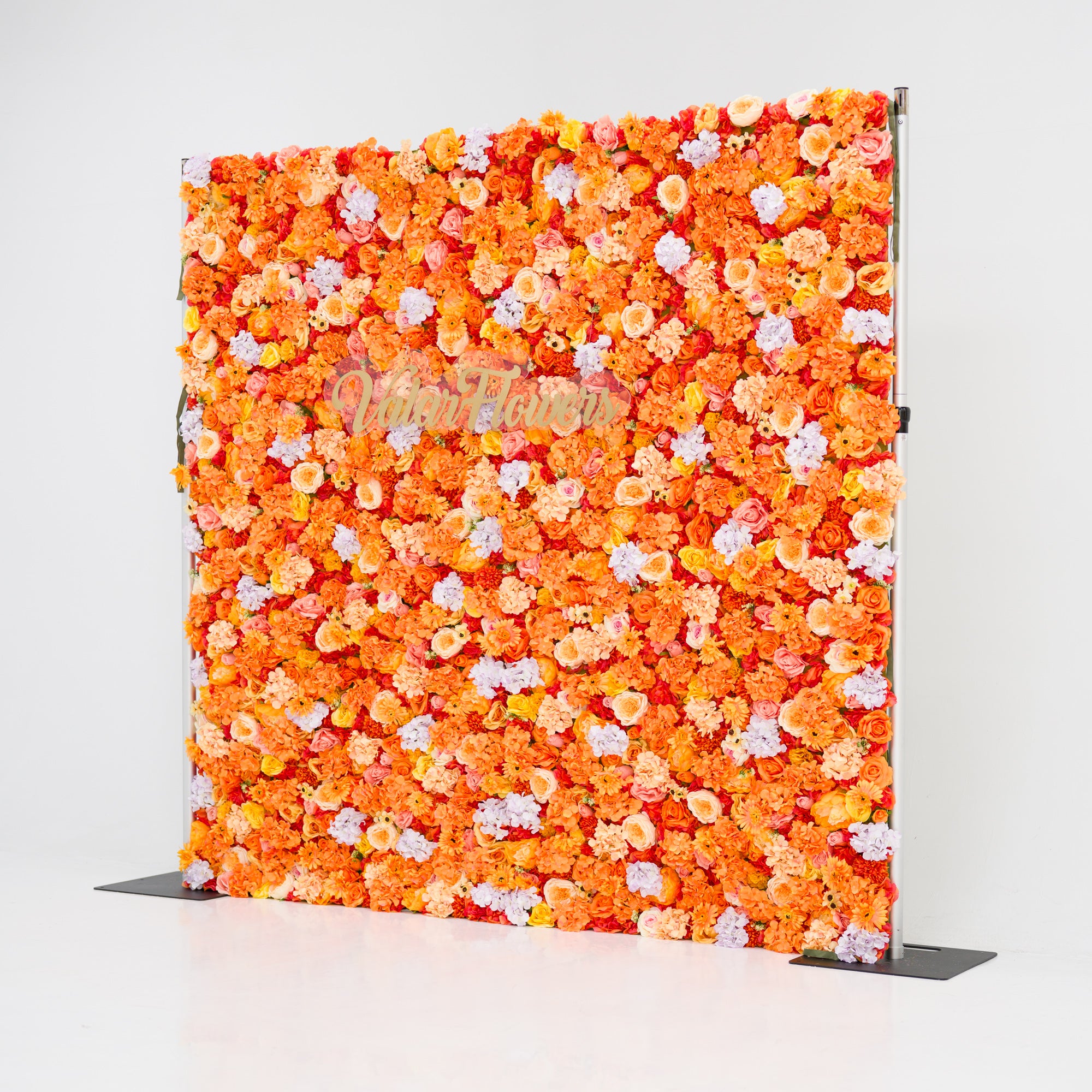 This image showcases a vibrant floral backdrop by Valar Flowers, featuring a dense arrangement of artificial flowers in warm shades of orange, peach, and hints of pink and purple. The textured display exudes energy, with the brand’s name elegantly integrated in gold. Set against a plain white indoor background, it’s perfect for adding a bold, nature-inspired element to events or interiors.