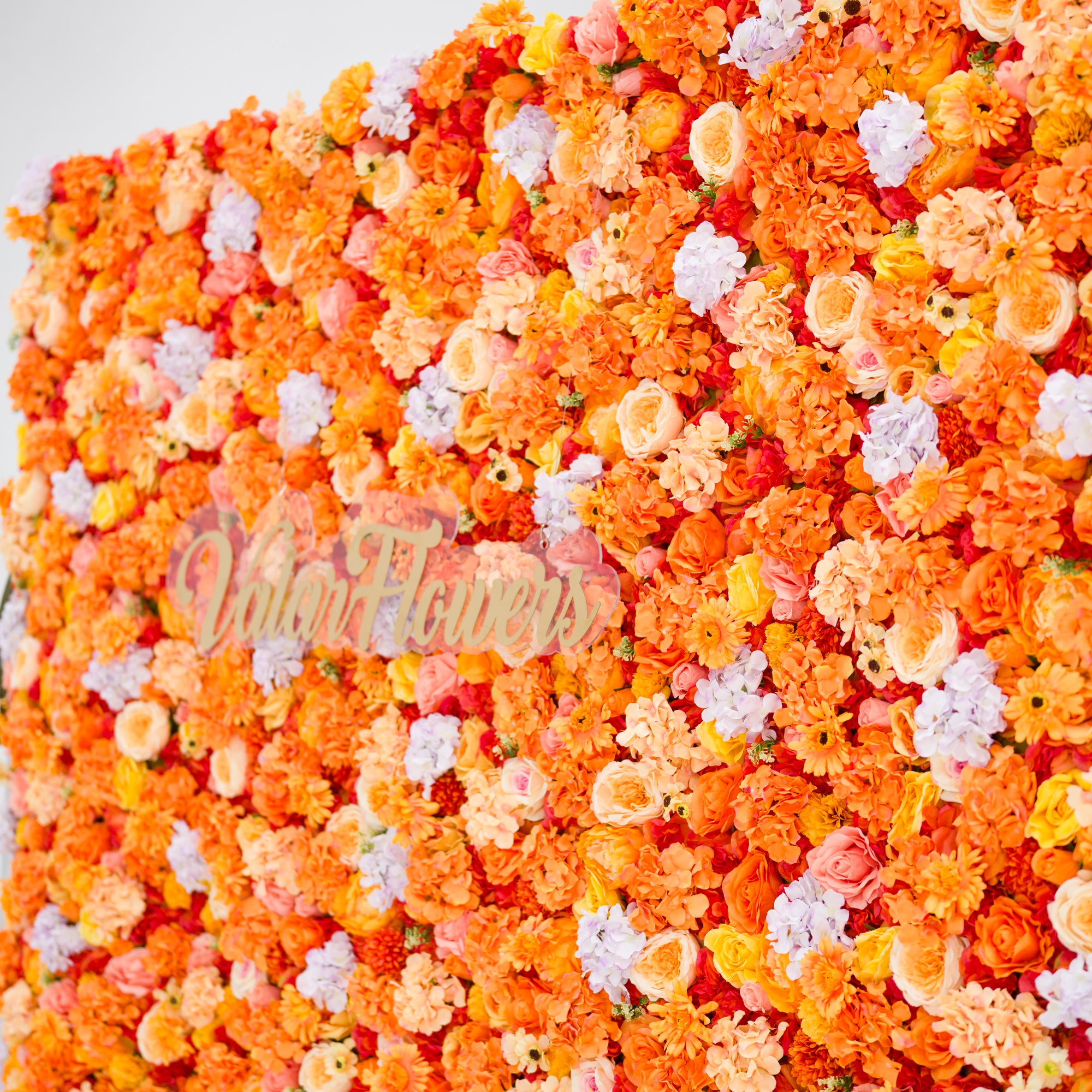 This image showcases a vibrant floral backdrop by Valar Flowers, featuring a dense arrangement of artificial flowers in warm shades of orange, peach, and hints of pink and purple. The textured display exudes energy, with the brand’s name elegantly integrated in gold. It’s perfect for adding a bold, nature-inspired element to events or interiors.