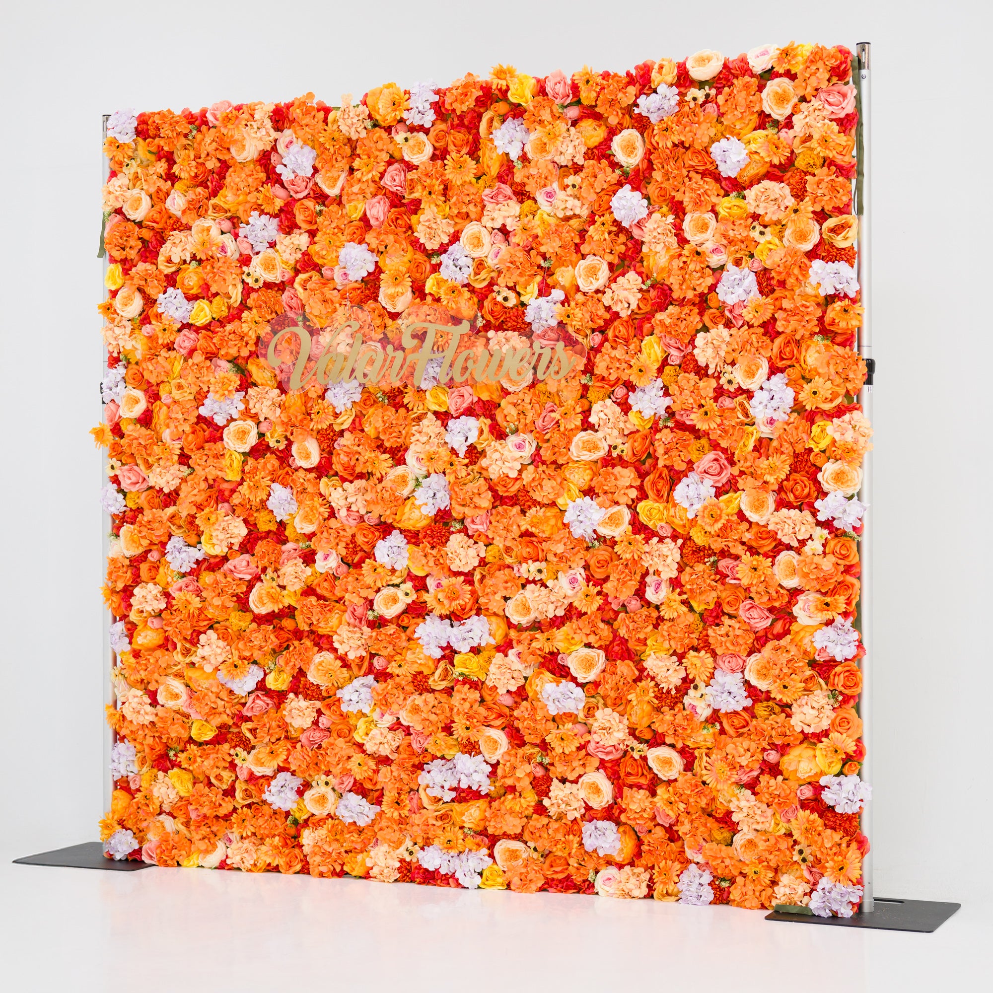 This image showcases a vibrant floral backdrop by Valar Flowers, featuring a dense arrangement of artificial flowers in warm shades of orange, peach, red, and hints of pink and purple. The textured display exudes energy, with the brand’s name elegantly integrated in gold. Set against a plain white indoor background, it’s perfect for adding a bold, nature-inspired element to events or interiors, highlighting Valar Flowers’ craftsmanship in eye-catching, sophisticated decor.