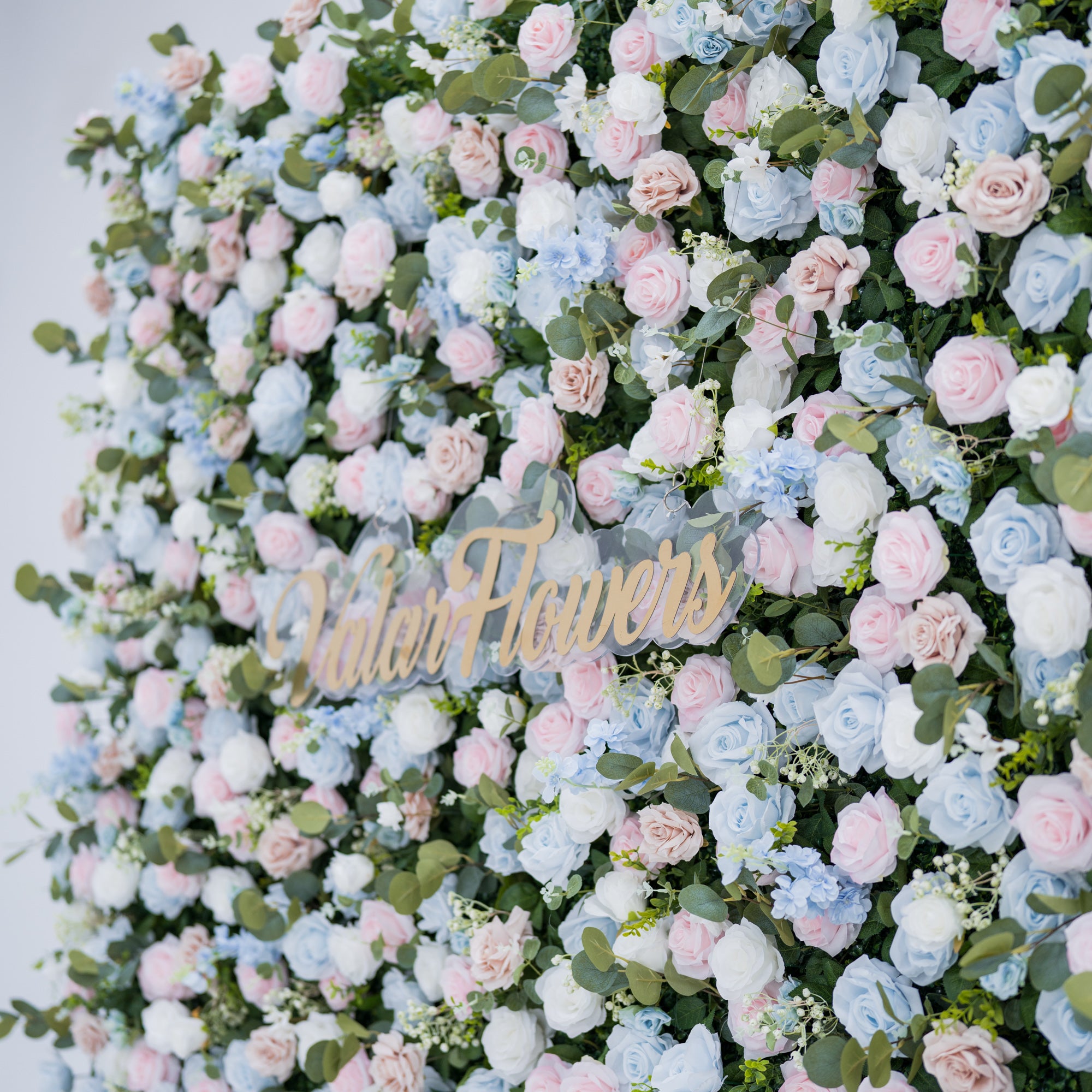 This image showcases a vibrant floral backdrop by Valar Flowers, featuring a dense arrangement of artificial flowers in shades of light blue, pink, white, and blush. Complemented by green foliage, the textured display exudes elegance. A gold “Valar Flowers” sign is integrated at the center, highlighting the brand’s craftsmanship in creating sophisticated, nature-inspired event decor.