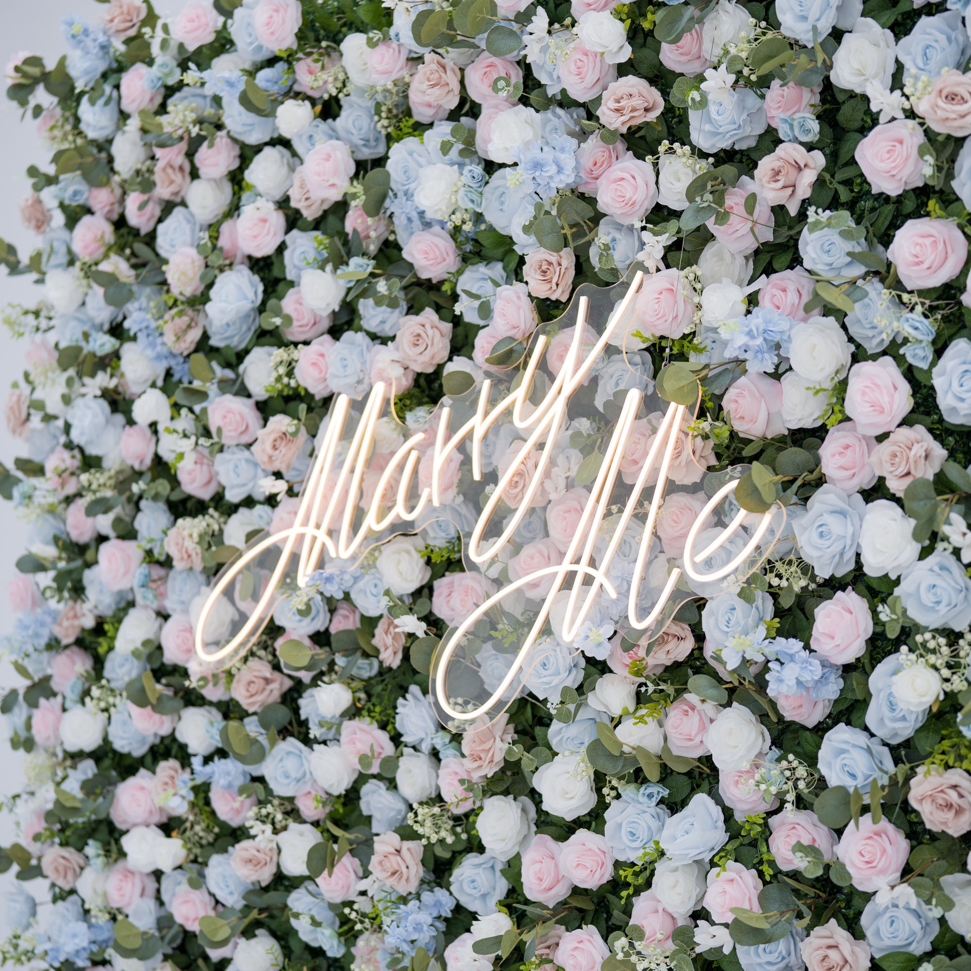 This image features a lush artificial floral backdrop with **roses** as the dominant flower type, in shades of light blue, pink, blush, and white. Accents include small blooms and green foliage like eucalyptus. At the center, a “Marry Me” neon sign adds romance. Ideal for proposals or weddings, it showcases meticulous craftsmanship in replicating realistic florals, blending various rose hues and textures for a sophisticated, eye-catching event decor piece.