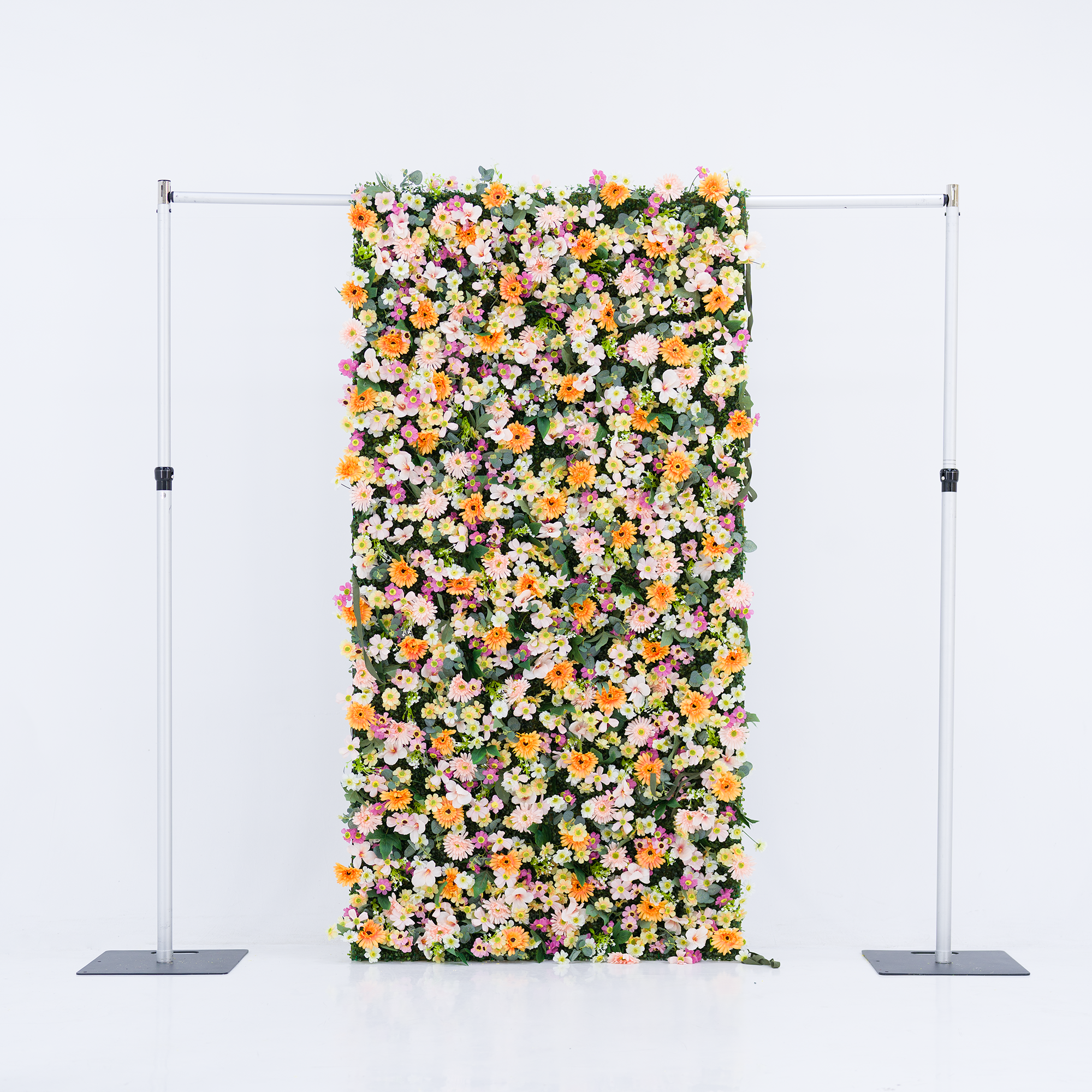 This vertical floral backdrop (mounted on a sleek silver frame, set against a plain white background) bursts with dense clusters of warm orange daisies, soft pale pink blooms, crisp white flowers, and subtle purple accents. Lush green foliage weaves through the vibrant petals, creating a full, cheerful display. The bright pastel-and-orange palette evokes a fresh, lively vibe—ideal for event backdrops, photo booths, or festive decor setups.
