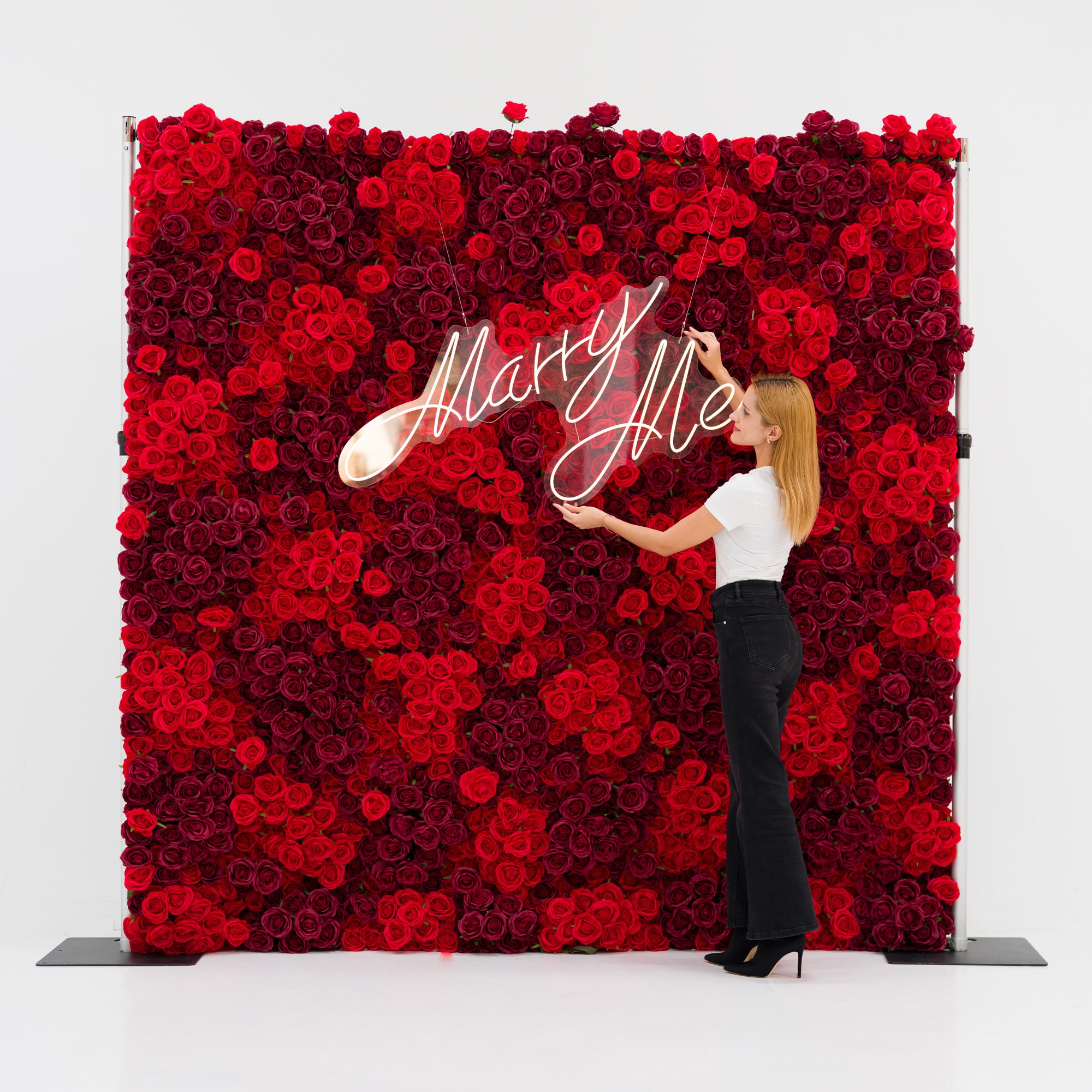 This striking backdrop (against a plain white background) is dense with layered red and burgundy roses, creating a lush, romantic texture. A woman (white top, black wide-leg pants, heels) adjusts a glowing "Marry Me" neon sign (cursive, light-lit) centered on the blooms. The bold, rich palette evokes passion and elegance, making it ideal for proposal setups, romantic events, or wedding decor.