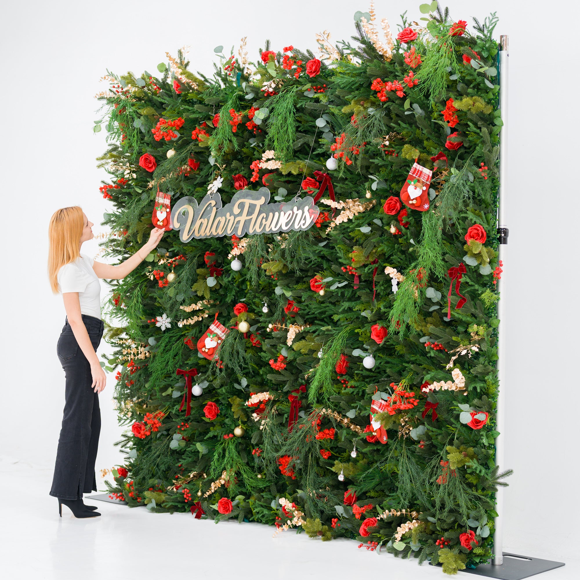This tall festive backdrop (against a plain white background) is lush with green foliage, red roses, clustered blooms, and red berries, accented by mini Santa stockings, gold/silver ornaments, and a polished “ValarFlowers” sign. A woman (white top, black wide-leg pants, heels) adjusts a stocking, adding a dynamic styling touch. The rich red-and-green palette blends cozy holiday charm with elegant texture—ideal for seasonal events or festive decor setups.