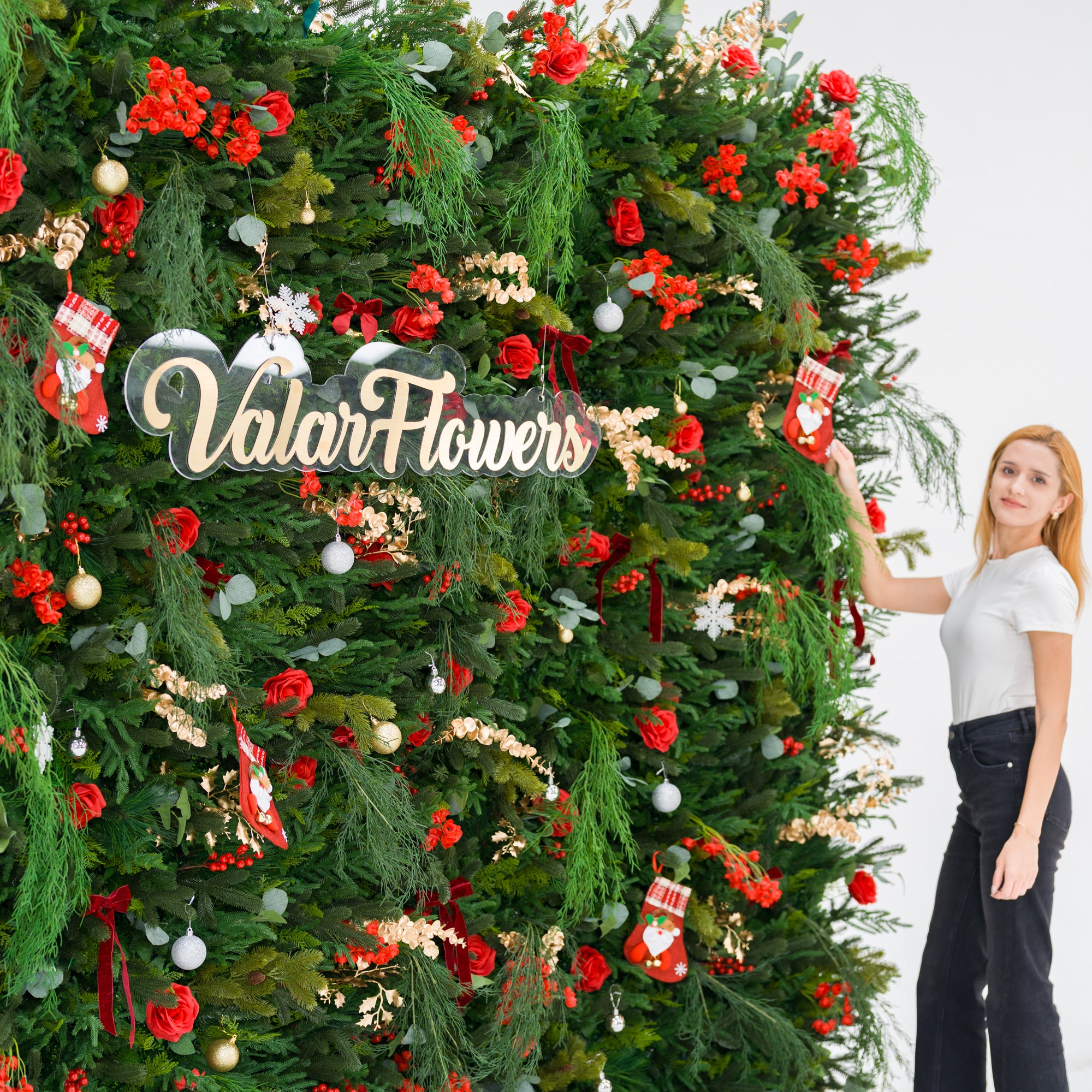 This lush festive greenery wall (against a plain white background) is dense with pine/feathery foliage, red roses, and red berry clusters. Accents include mini Santa stockings, gold/silver ornaments, and gold sprays, plus a polished “ValarFlowers” sign. A woman (white top, black jeans) adjusts a stocking, adding a dynamic styling touch. The rich red-and-green palette blends cozy holiday charm with elegant texture—ideal for seasonal events or festive decor setups.