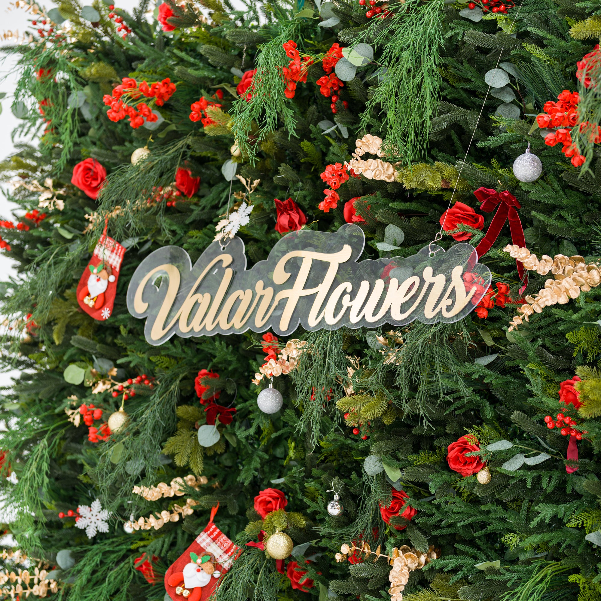 This close-up captures a lush festive arrangement: dense green foliage (pine, feathery fronds) mixed with bold red roses, clustered blooms, and red berries. Accents include mini Santa stockings, gold/silver ornaments, snowflakes, and a polished “ValarFlowers” sign. Varied textures—soft petals, lush greenery, shiny decor—blend with the rich red-and-green palette, evoking cozy holiday charm, perfect for seasonal decor or festive event styling.