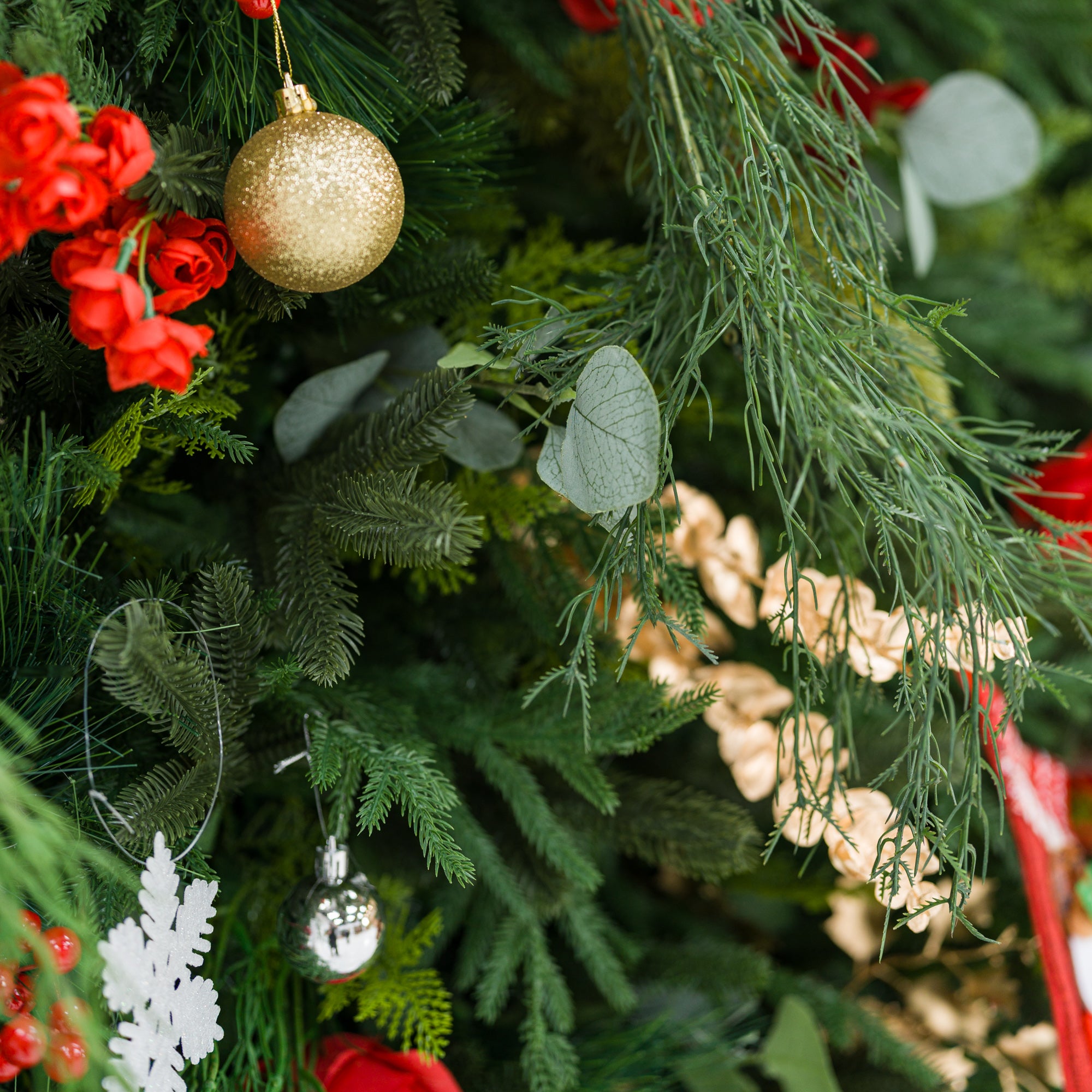 This detailed close-up captures a festive arrangement: dense green foliage (pine, feathery fronds) mixed with bold red blooms, plus accents like a glittering gold ornament, a shiny silver bauble, a white snowflake decor, and delicate gold sprays. Soft eucalyptus leaves add texture, while the rich red-and-green palette (paired with metallic touches) evokes cozy holiday charm—ideal for seasonal decor details or festive styling inspiration.