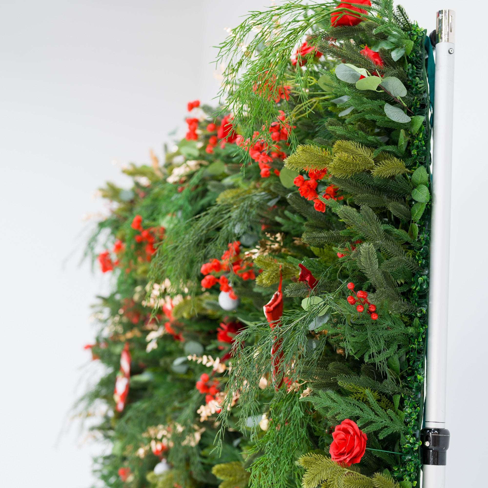 This angled view of a festive backdrop (against a plain white background) features dense green foliage (pine, feathery fronds) mixed with bold red roses, clustered blooms, and red berries. Accents include mini Santa stockings, a silver ornament, and delicate gold sprays. A metallic frame edge is visible, grounding the lush, layered design. The rich red-and-green palette evokes cozy holiday charm—ideal for seasonal event decor or Christmas-themed backdrops.