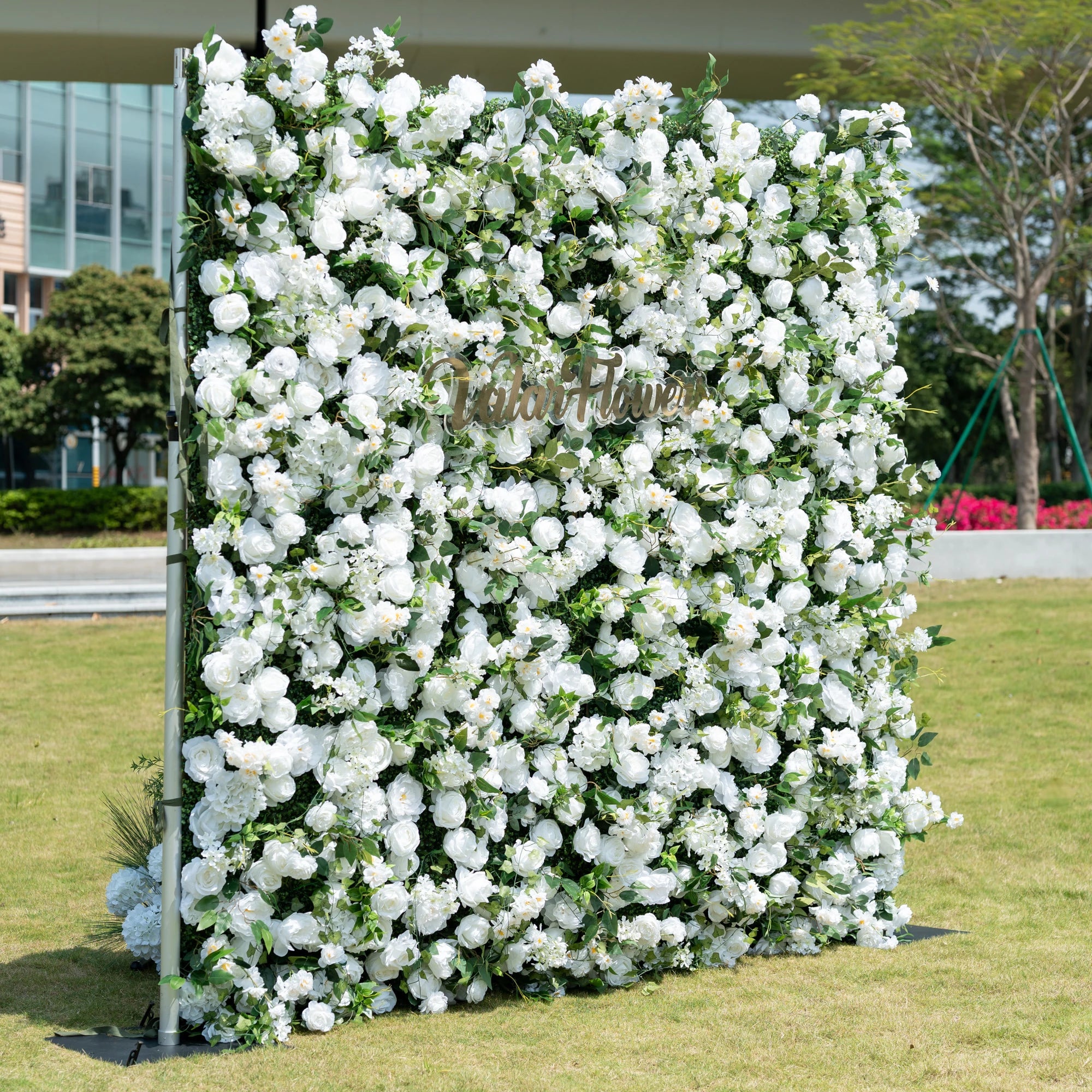 This is a premium all-white artificial flower wall backdrop by Valar Flowers, featuring a lush, dense design of soft roses, peonies, delicate baby’s breath, and subtle green foliage. Centered with the metallic "ValarFlowers" logo, this reusable, maintenance-free display creates an elegant, timeless focal point. Ideal for outdoor weddings, photo booths, luxury events, and brand activations, it offers a clean, romantic aesthetic perfect for professional styling.