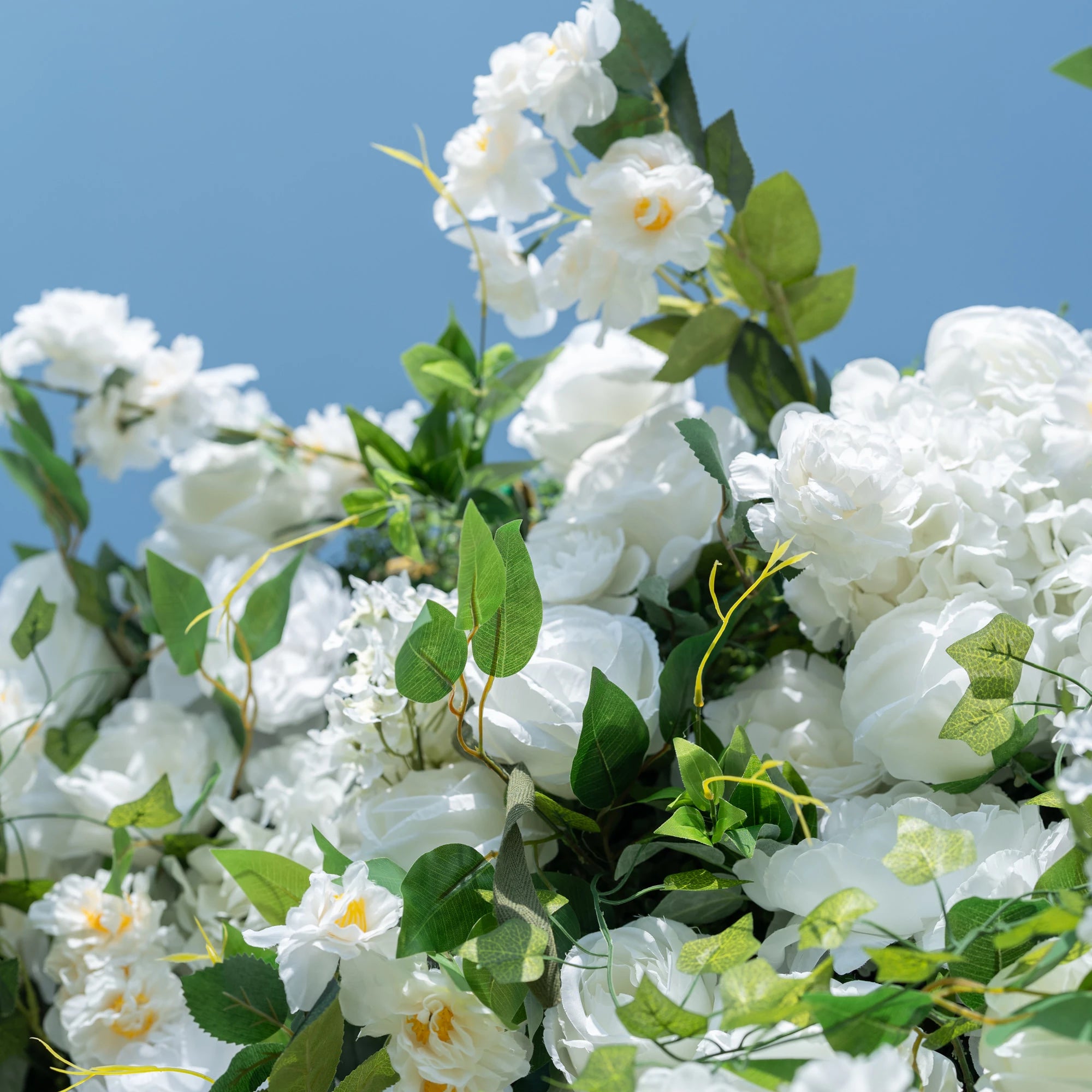 This close-up highlights the premium craftsmanship of Valar Flowers’ artificial floral arrangement, featuring lush white roses, peonies, and delicate filler blooms paired with vibrant, realistic green foliage. The soft-textured petals mimic fresh flowers, while layered leaves and subtle yellow accents create a lifelike, maintenance-free display. Ideal for weddings, photo booths, and luxury events, this durable decor offers a clean, romantic aesthetic perfect for professional styling.