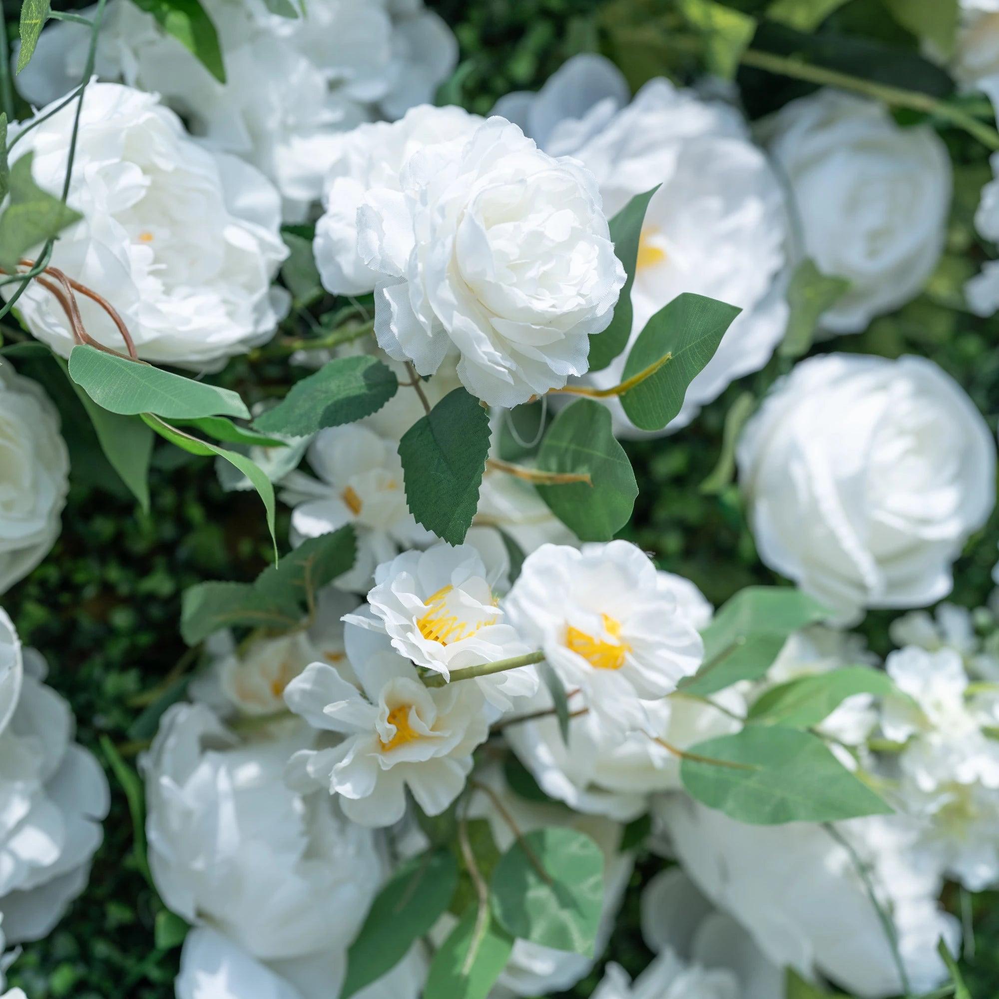 This close-up showcases the premium details of Valar Flowers’ artificial white floral wall, featuring lush, hyper-realistic roses, peonies, and delicate yellow-centered filler blooms paired with vibrant green foliage. Soft-textured petals mimic fresh flowers, while layered leaves and stems create a lifelike, maintenance-free display. Ideal for weddings, photo booths, and luxury events, this durable decor offers a clean, romantic aesthetic perfect for professional styling.