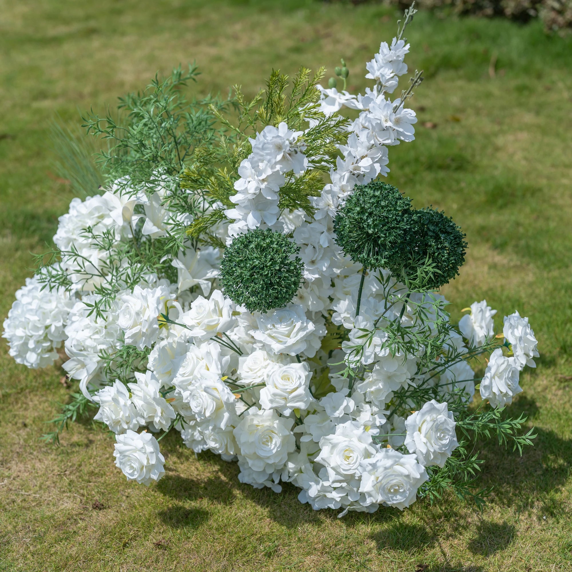This premium artificial floral arrangement by Valar Flowers features a lush mix of white roses, hydrangeas, lilies, delphiniums, and textured green foliage, including spherical green accents. Designed for weddings, photo booths, and luxury events, this dense, realistic display offers a clean, timeless aesthetic. Maintenance-free and durable, it brings a romantic, elegant touch to any setting, perfect for professional styling and event decor.