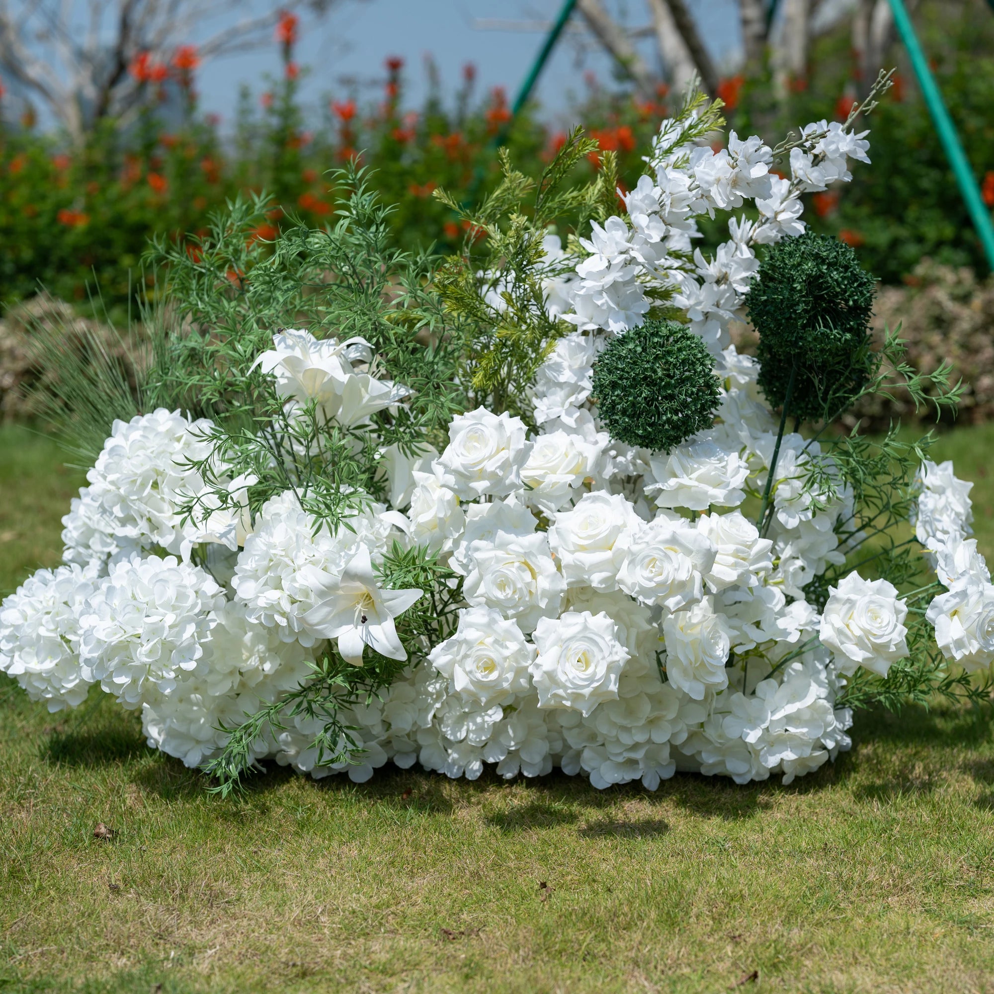This full view showcases Valar Flowers’ premium artificial floral arrangement, featuring a lush mix of white roses, hydrangeas, lilies, delphiniums, and textured green foliage including spherical accents. Set against an outdoor garden backdrop, the dense, realistic display mimics fresh blooms, offering a clean, timeless aesthetic. Maintenance-free and durable, this elegant piece is ideal for weddings, photo booths, and luxury events, perfect for professional styling.
