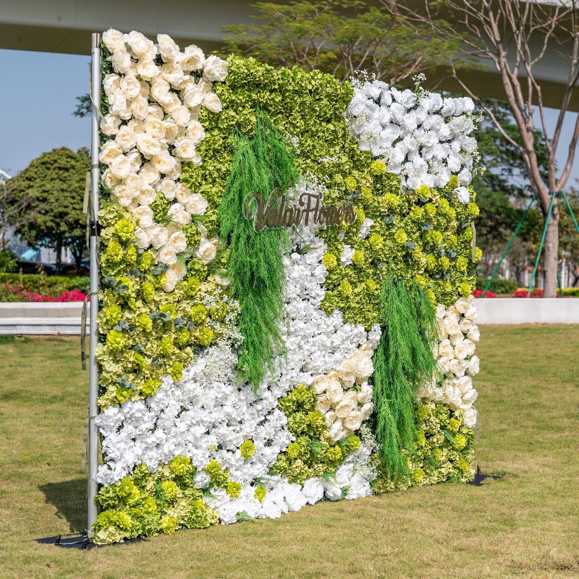 This premium artificial flower wall backdrop by Valar Flowers features a lush, nature-inspired design with vibrant green hydrangeas, crisp white blooms, soft ivory roses, and cascading green foliage, accented by the custom "ValarFlowers" logo. Reusable and maintenance-free, it creates a fresh, elegant focal point ideal for outdoor weddings, garden parties, photo booths, and luxury events, offering long-lasting beauty for professional decor.