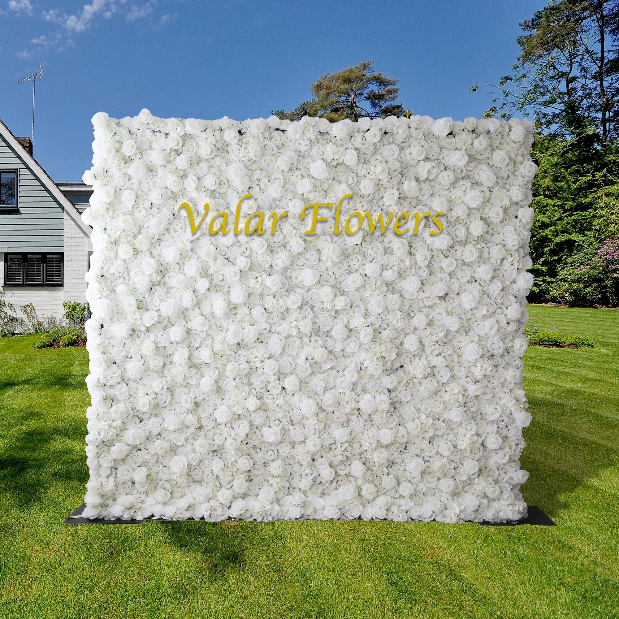 A large white artificial flower wall by Valar Flowers set in an outdoor location with a clear blue sky and a house visible in the background.
