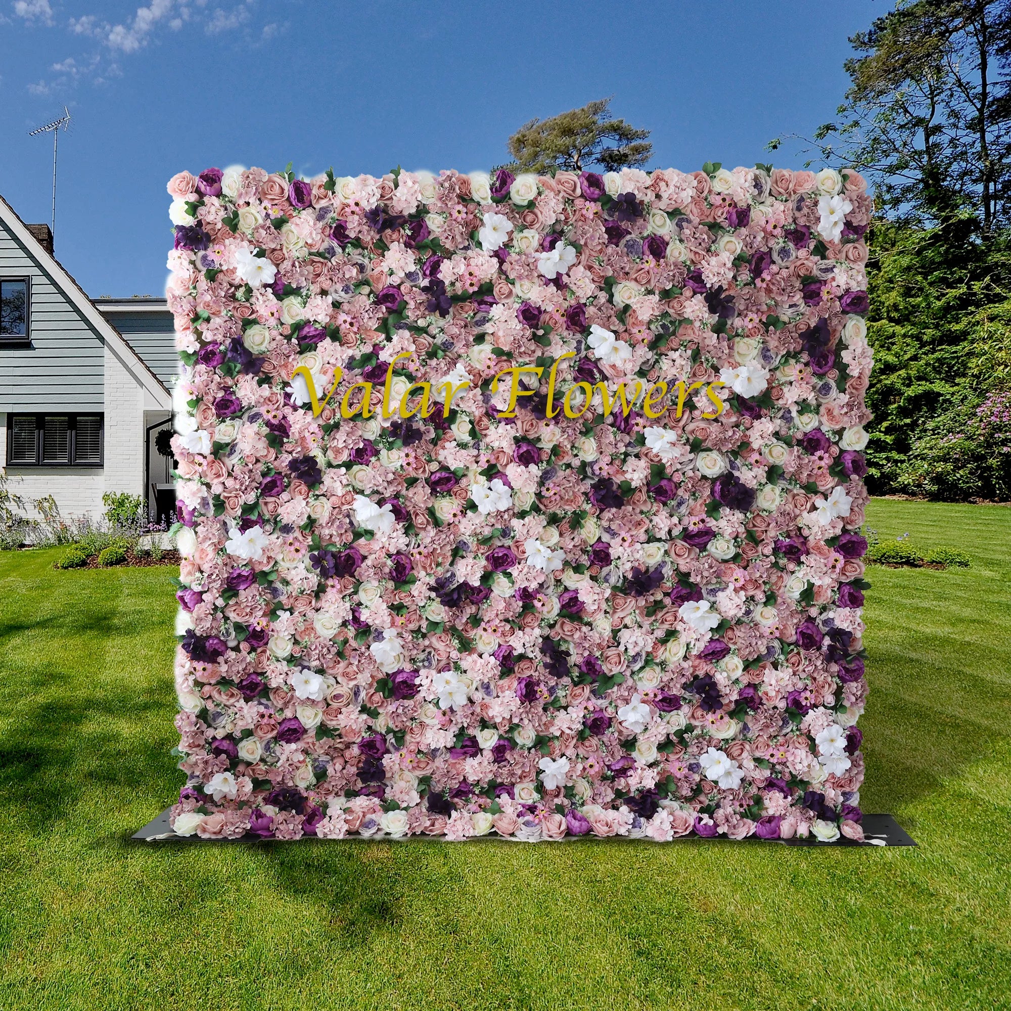 This romantic outdoor floral backdrop features a dense, layered arrangement of artificial blooms in a lush palette of blush, dusty rose, deep plum, and soft white. A sleek, glossy gold “Valar Flowers” logo sits at its center, adding a refined touch. Set on a sunlit green lawn, with a charming cottage and trees in the background, it exudes vintage elegance and warmth, making it a stunning focal point for weddings, photoshoots, or upscale events.