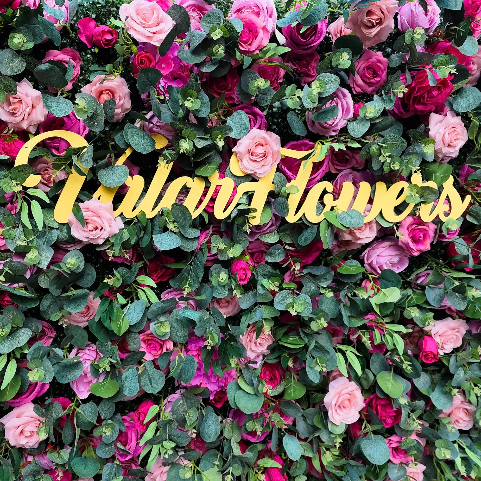 This image showcases a lush floral wall with roses in shades of pink, red, and purple, interspersed with green eucalyptus leaves. Prominently displayed is golden cursive text reading “Valar Flowers”. The vibrant blend of colors and textures creates a visually appealing, elegant scene, ideal for decorative purposes in events or as an artistic floral display.