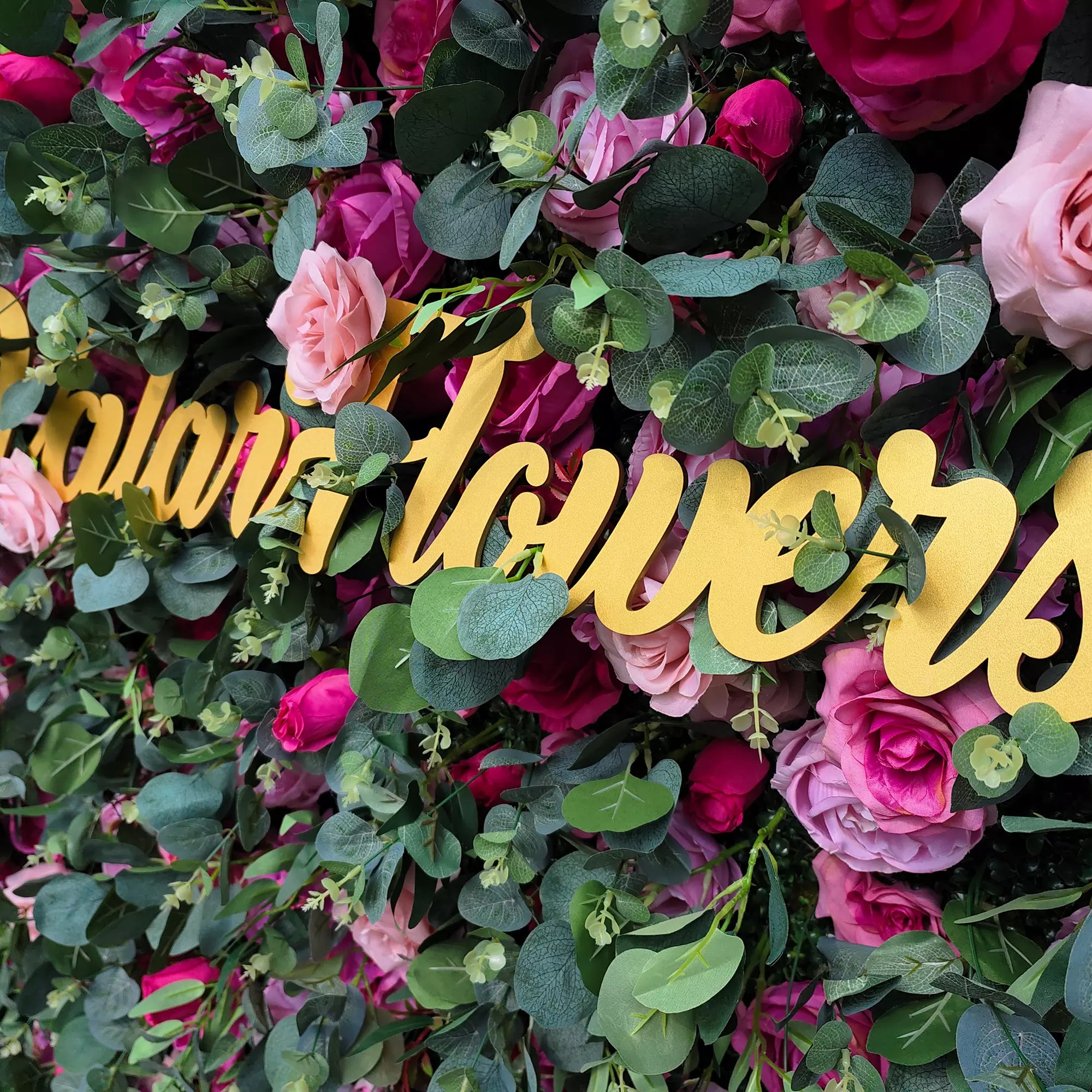 This close - up image showcases a lush floral arrangement with roses in shades of pink and magenta, interspersed with green eucalyptus leaves. Prominently displayed is golden cursive text, partially reading “Valar Flowers”. The vibrant blend of colors and textures creates a visually appealing, elegant scene, ideal for decorative purposes in events or as an artistic floral display.