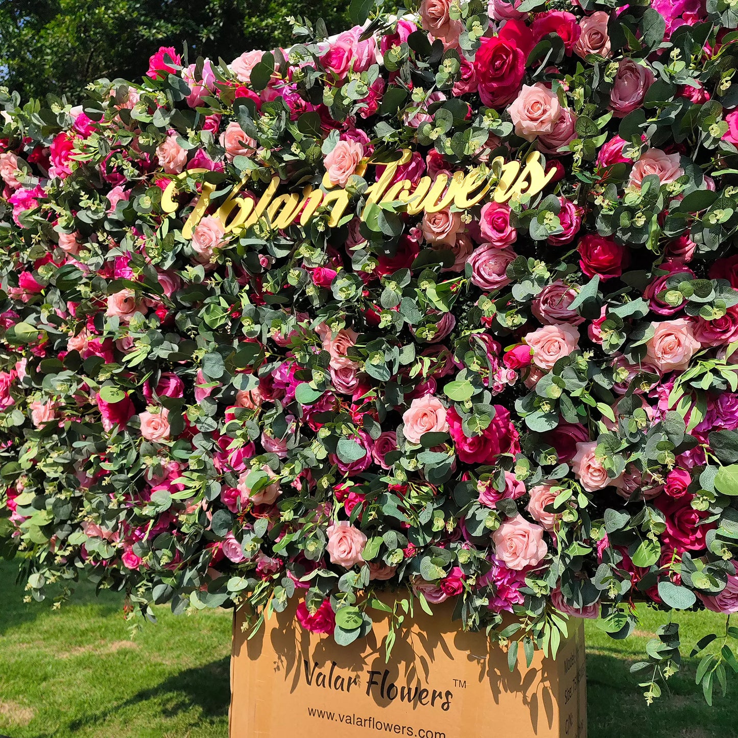 This image showcases a large, vibrant floral arrangement by Valar Flowers. It's densely packed with roses in shades of pink, red, and purple, interspersed with lush green eucalyptus leaves. A golden "Valar Flowers" sign is prominently displayed. The arrangement sits atop a cardboard box branded with "Valar Flowers" and is set outdoors on a grassy area, creating a visually striking and elegant display.