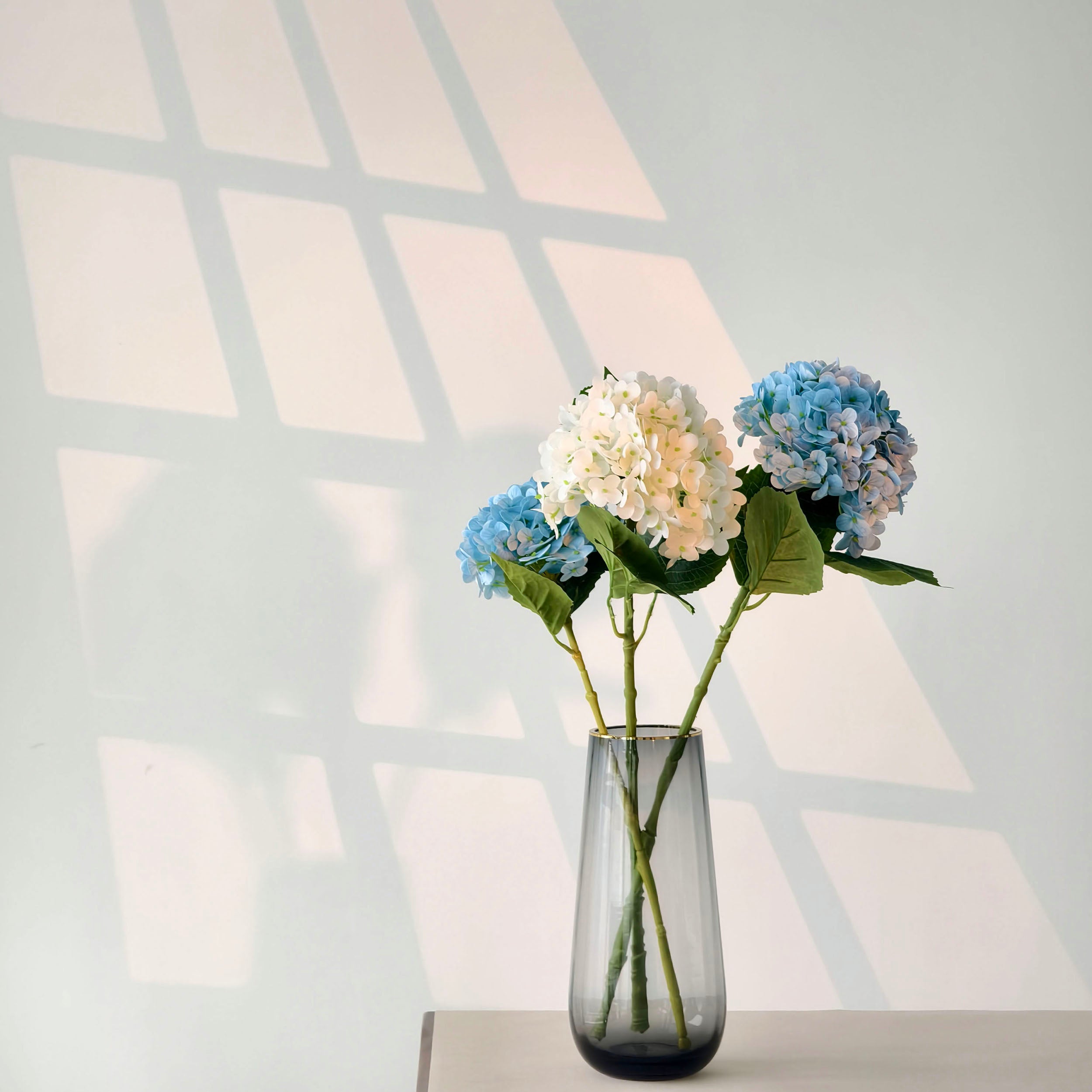 This arrangement has three hydrangeas—two soft blue, one white - pink gradient—with green leaves. Placed in a sleek, gradient - colored glass vase, it sits on a light table against a wall with grid - like shadows. The pastel hues and minimalist setup create a calm, elegant, and modern decor vibe.