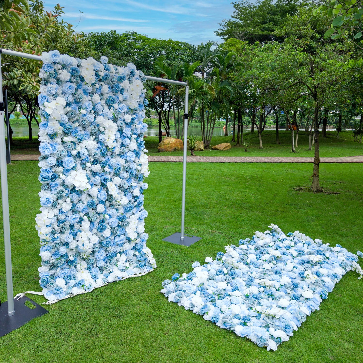 This photo depicts a serene outdoor setting featuring two large floral arrangements made of blue and white roses and white orchids. One arrangement is mounted on a metal frame, standing upright, while the other lies flat on the grass. Set against a backdrop of lush greenery, a body of water, and a clear sky, the scene exudes tranquility and elegance, ideal for events like weddings or garden parties.