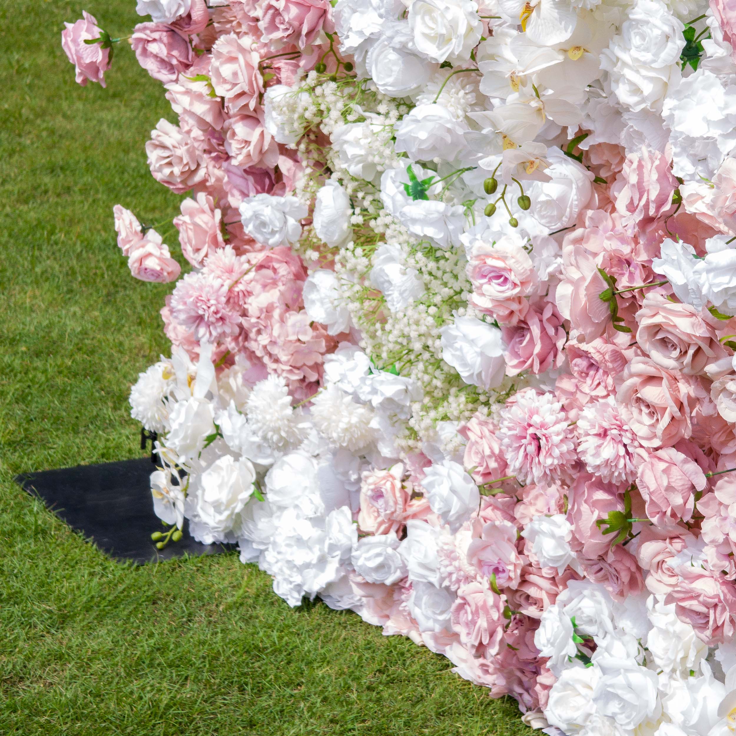 This backdrop is a lush floral wall made of artificial flowers, set against a grassy surface. It features a mix of white and various shades of pink roses, white chrysanthemums, and clusters of baby&